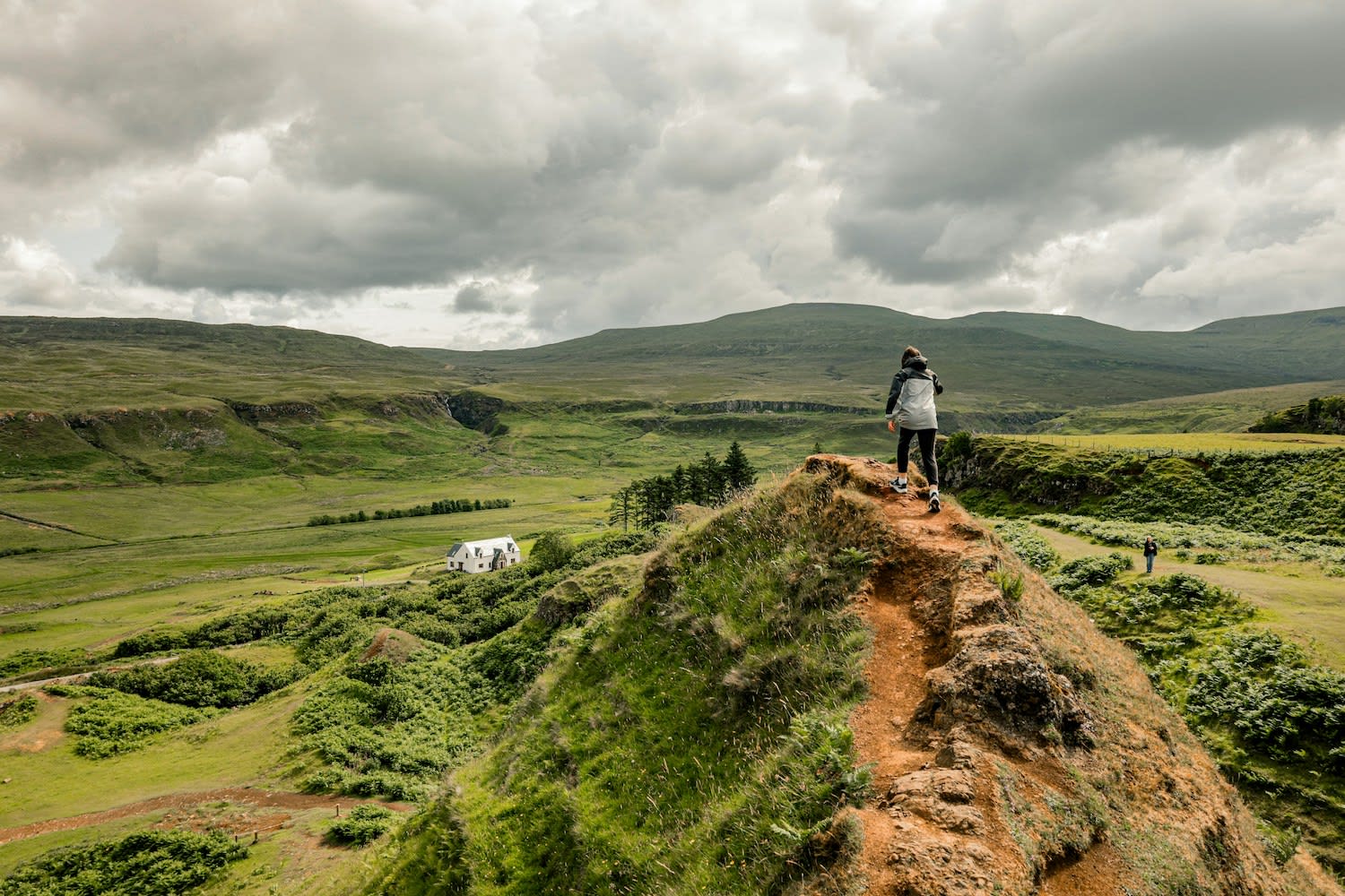 Persona caminando por una cresta de tierra en un paisaje montañoso y verde bajo un cielo nub
