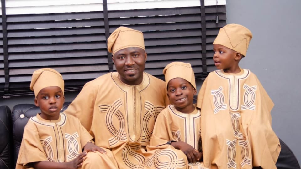 A father and his three young sons seated together in matching gold agbada and fila