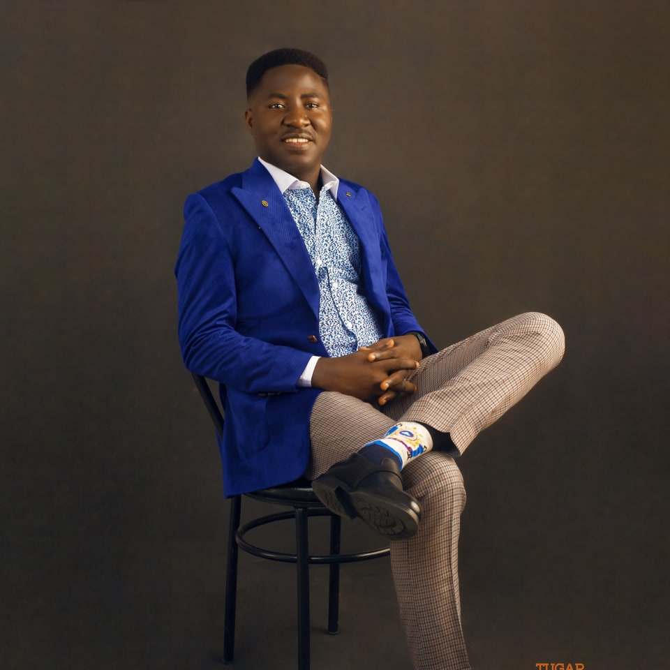 Man seated on a chair in a royal blue blazer and check trousers against a dark backdrop