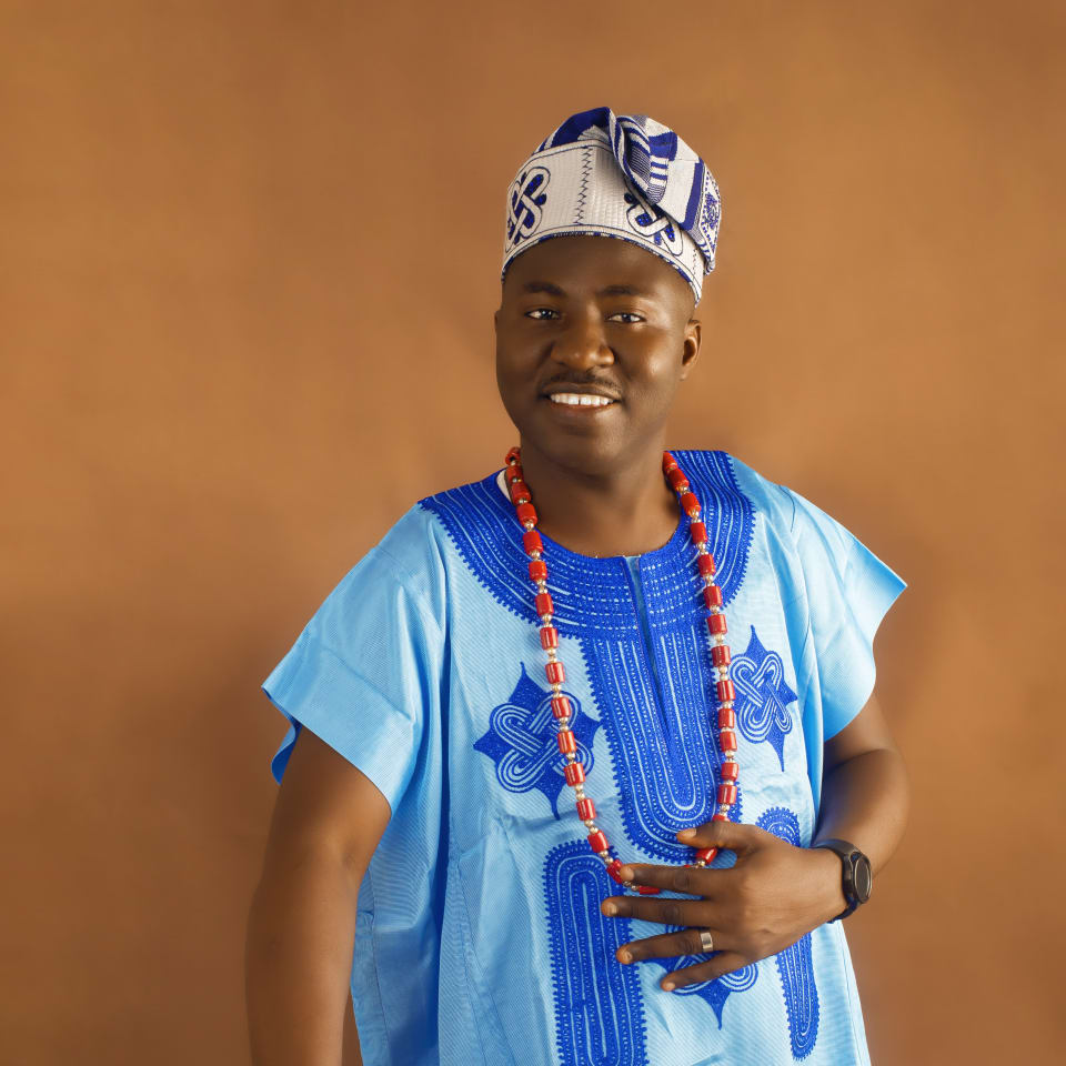 Smiling man in a sky blue buba with royal blue embroidery, fila and coral beads
