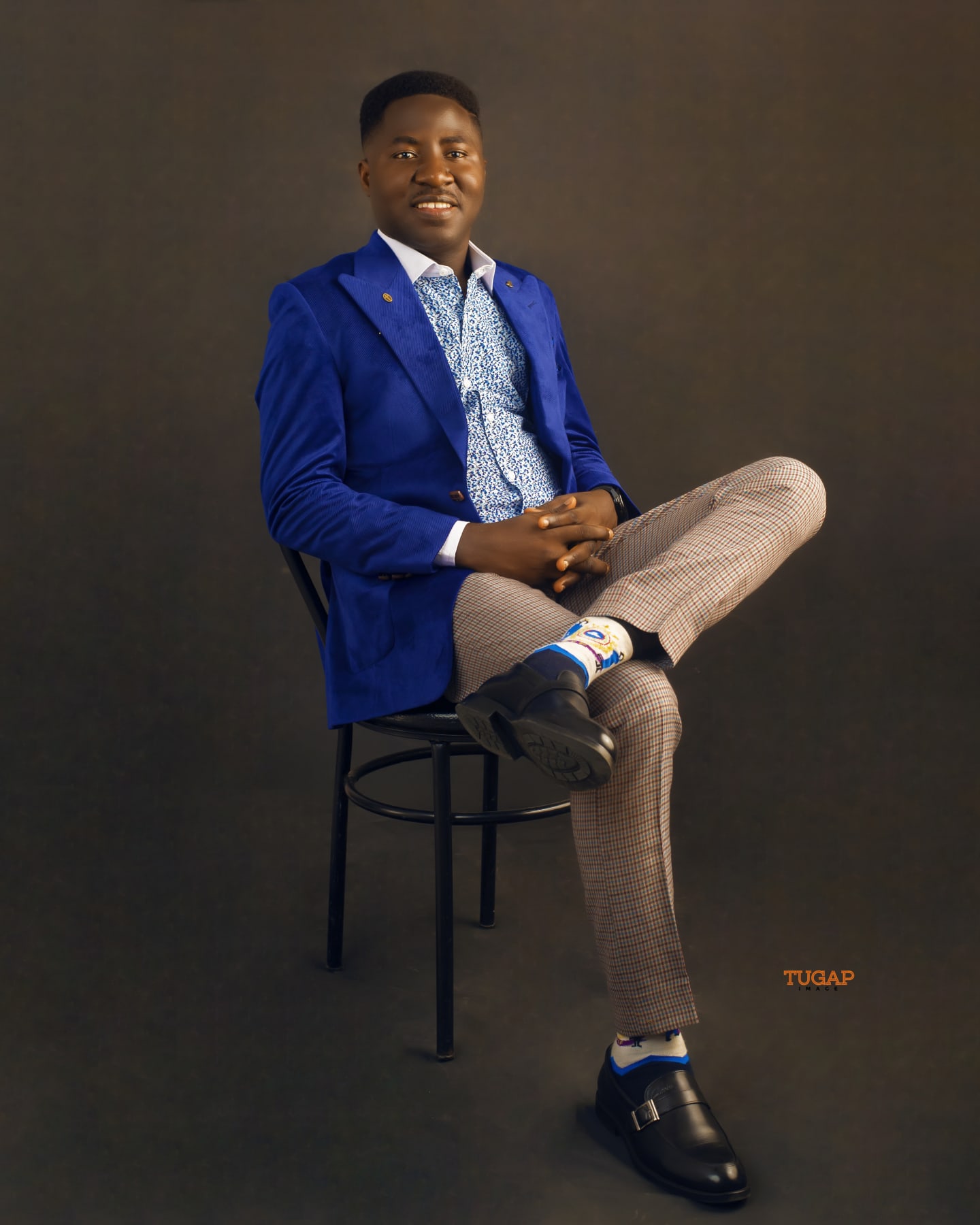 Man seated on a chair in a royal blue blazer and check trousers against a dark backdrop