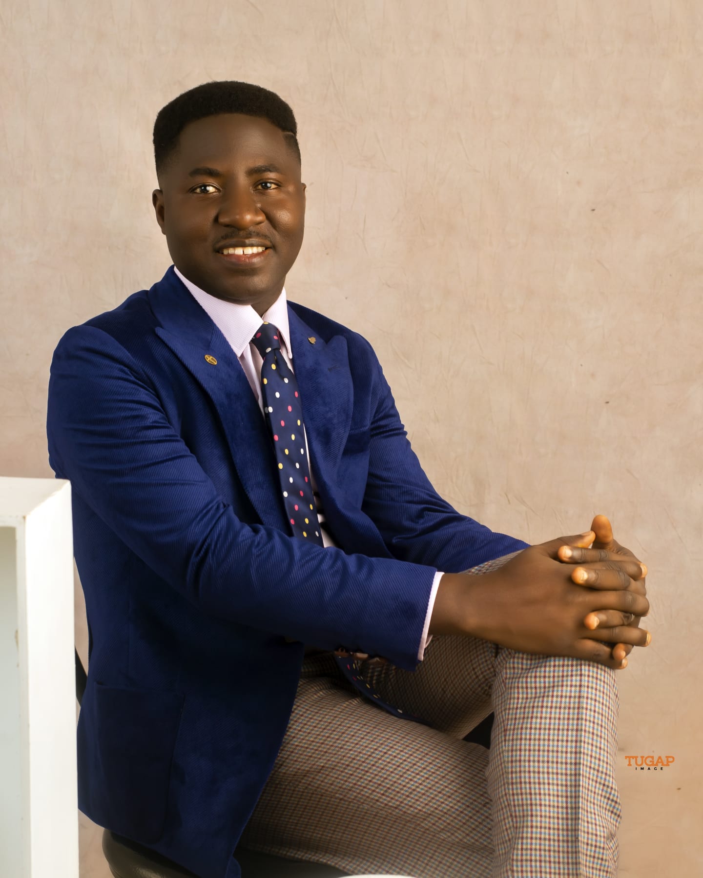 Smiling man seated in a tailored royal blue blazer, dotted tie and check trousers