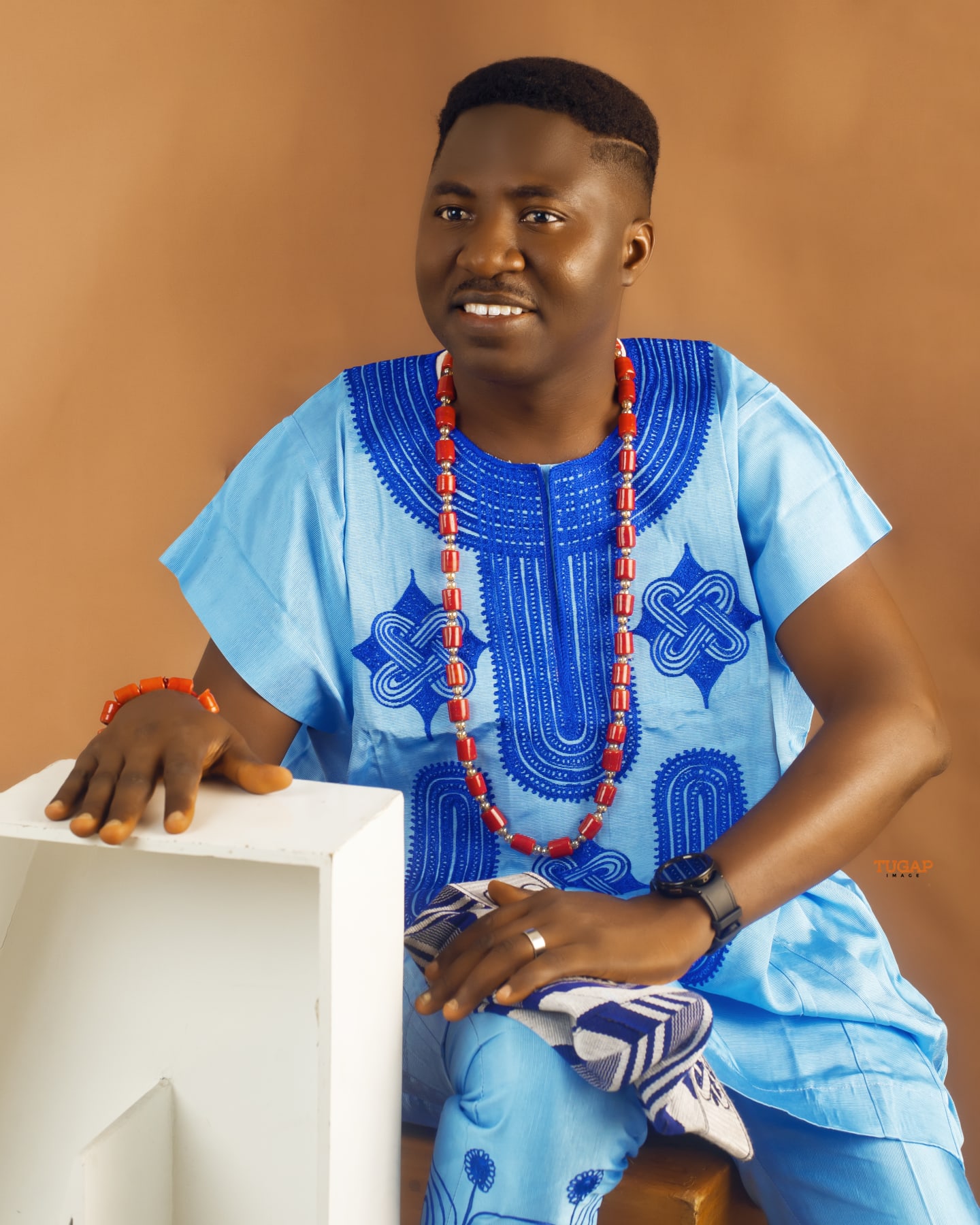 Man seated in a sky blue native outfit with bold royal blue embroidery and coral beads