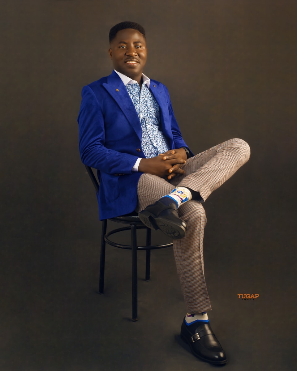 Man seated on a chair in a royal blue blazer and check trousers against a dark backdrop