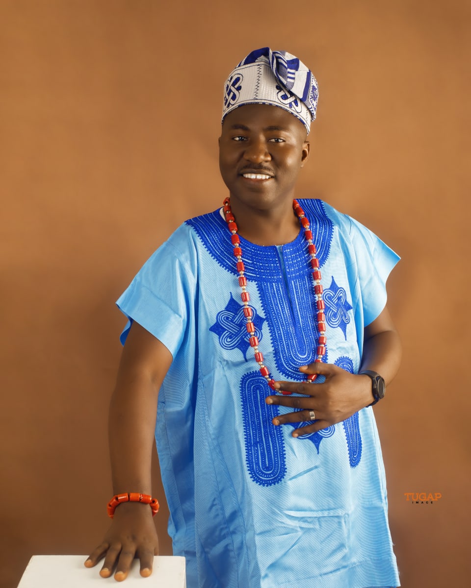 Smiling man in a sky blue buba with royal blue embroidery, fila and coral beads