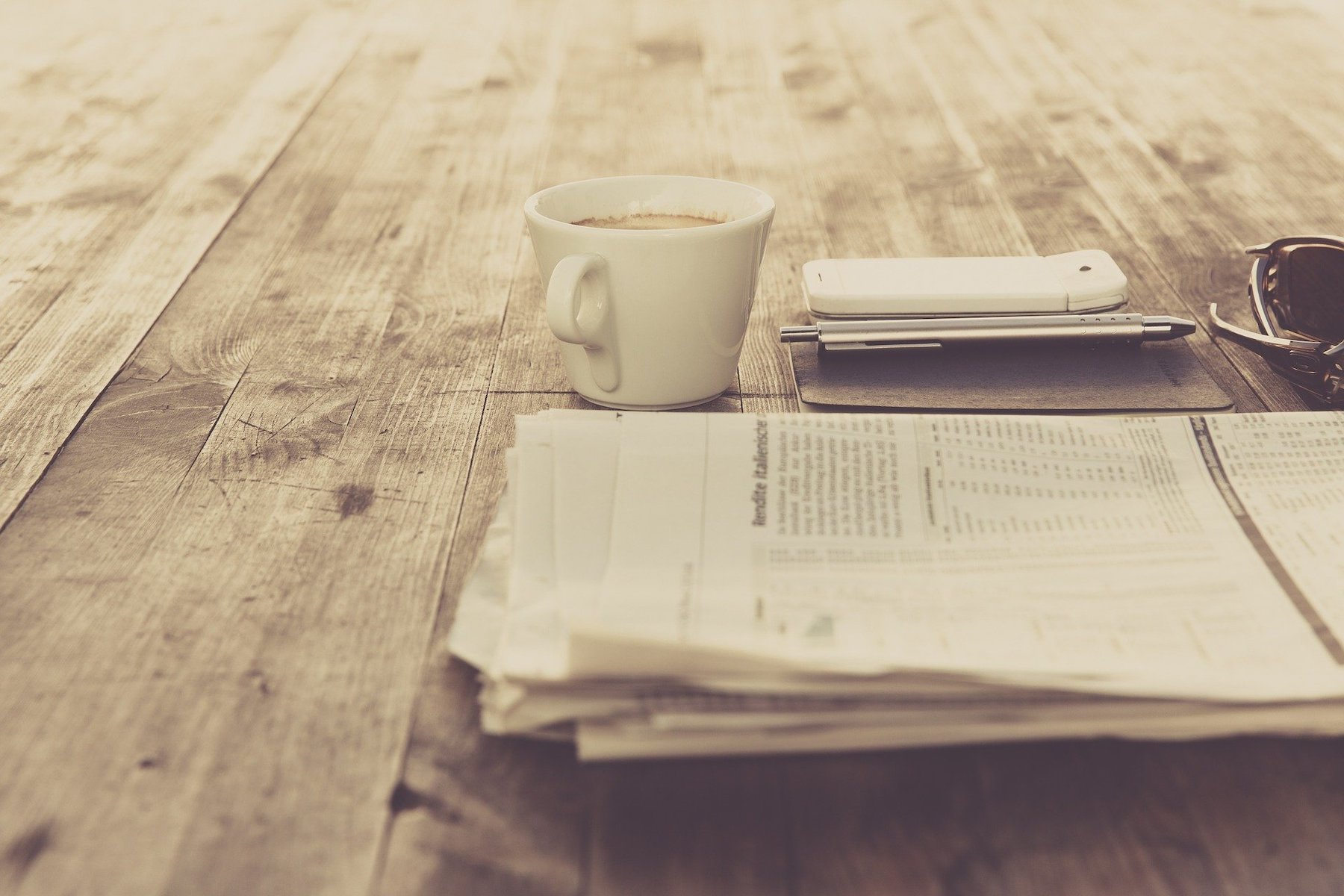 A coffee, phone, notebook and newspaper on a table.