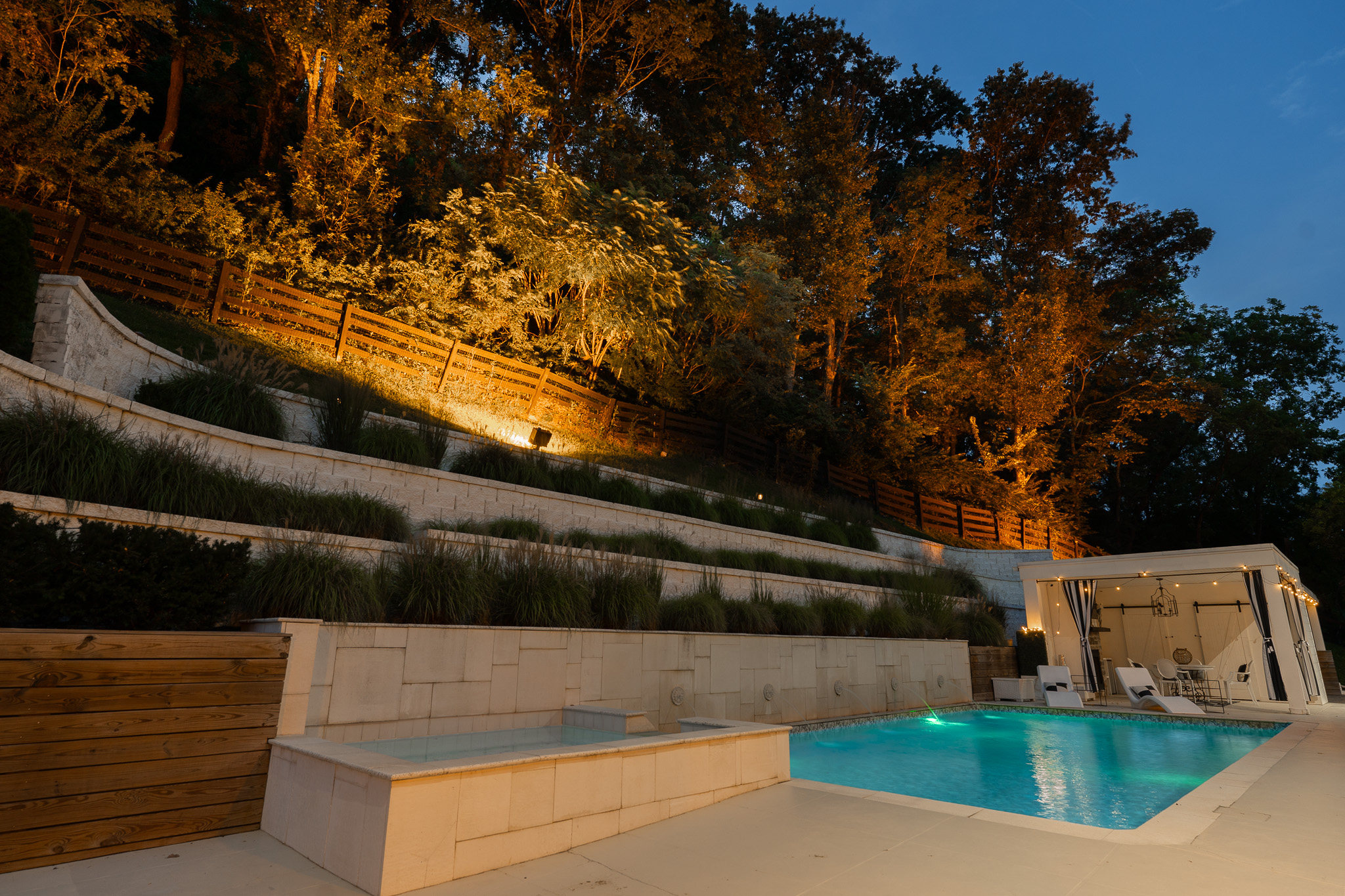The pool terrace at blue hour: the water lit turquoise, the limestone terraces washed from below, the wooded hillside uplit amber, and strung lights across the cabana.