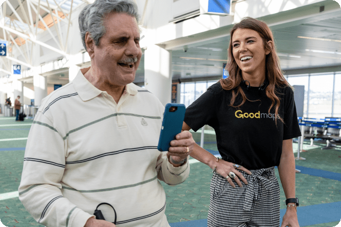 A blind man uses the GoodMaps app at an airport with a happy GoodMaps employee standing nearby