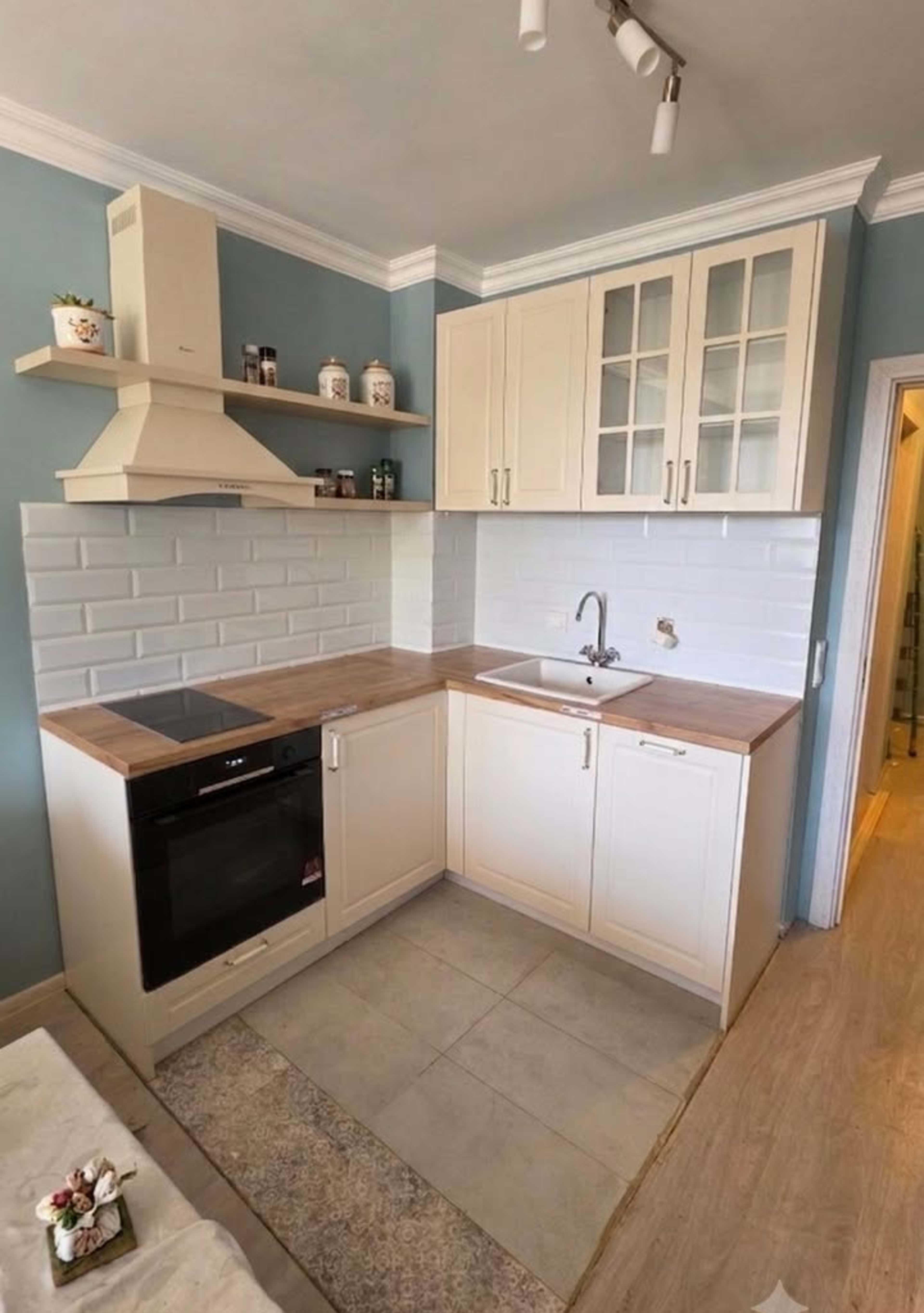 Cream L-shaped kitchen with shaker-style fronts, glazed doors, a wood-grain worktop and white brick-effect tiles.