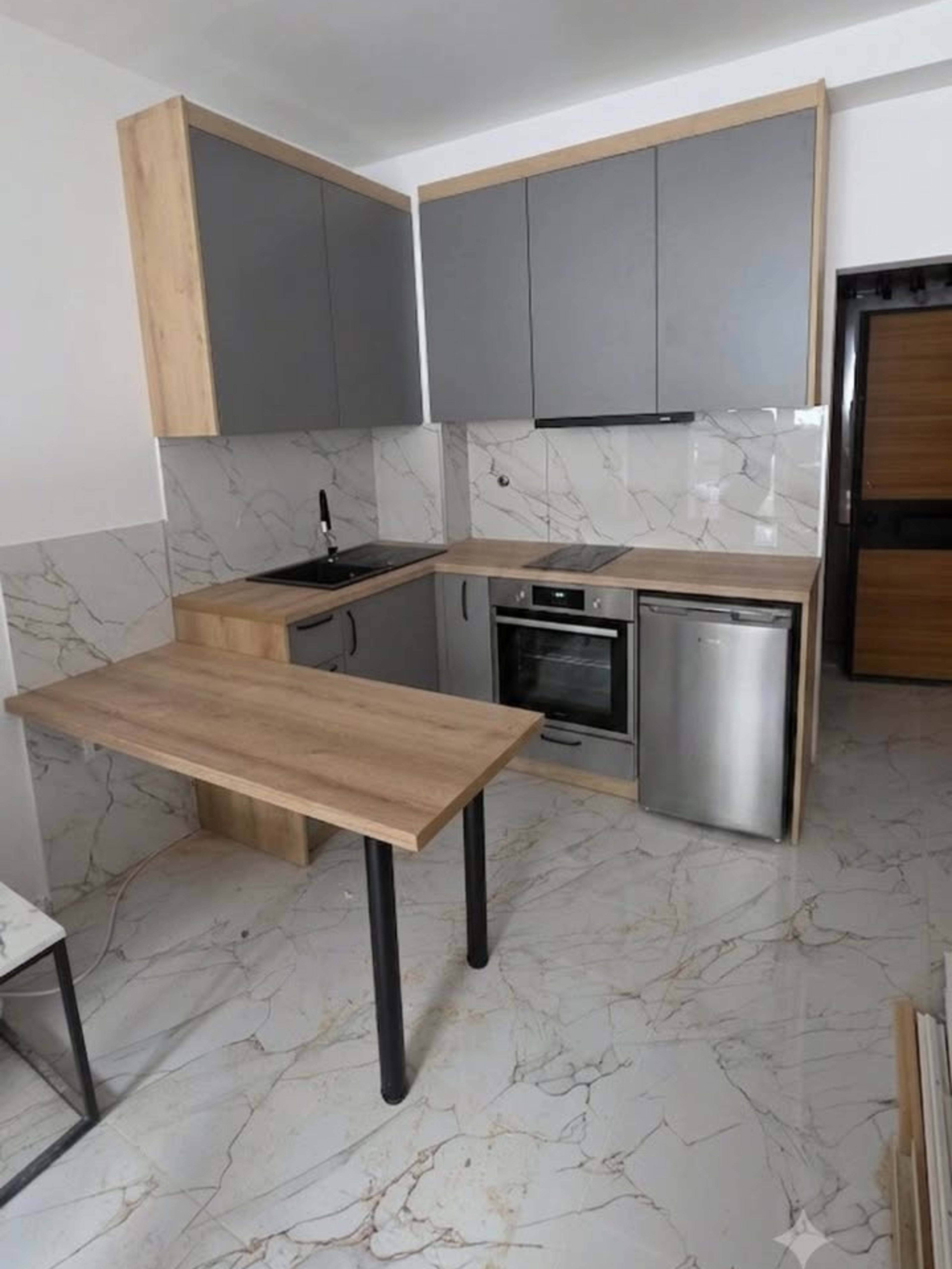 L-shaped kitchen with grey fronts in a wood-grain frame, a marble-effect splashback and a bar top on two black legs.
