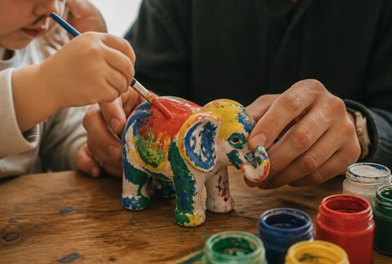 Dreijähriges Kind bemalt mit Hilfe eine weiße Elefantenfigur mit dicken bunten Pinselstrichen
