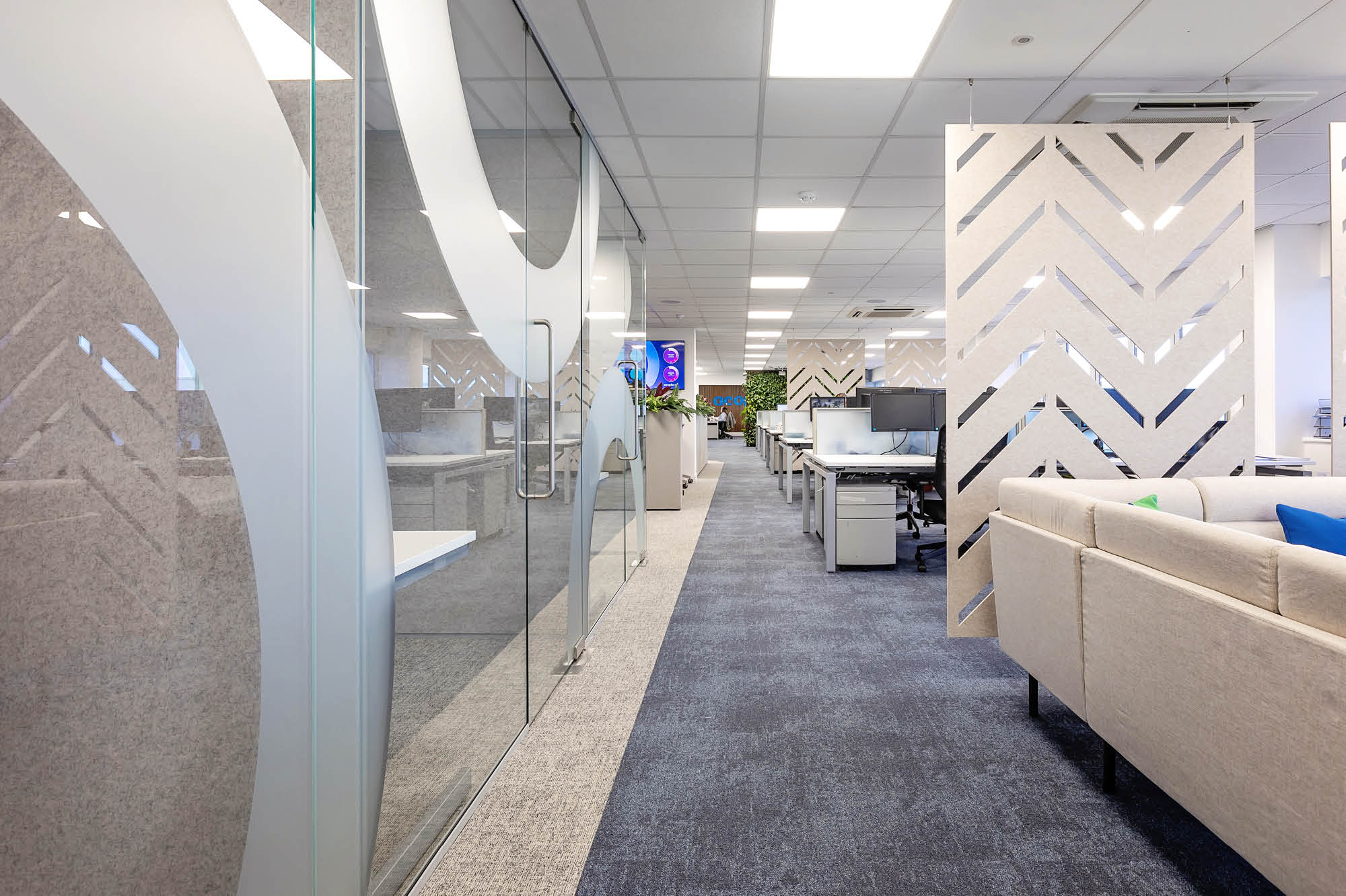 Modern office space with glass partitions and chevron-patterned dividers. The hallway leads to an open area with workstations and chairs.