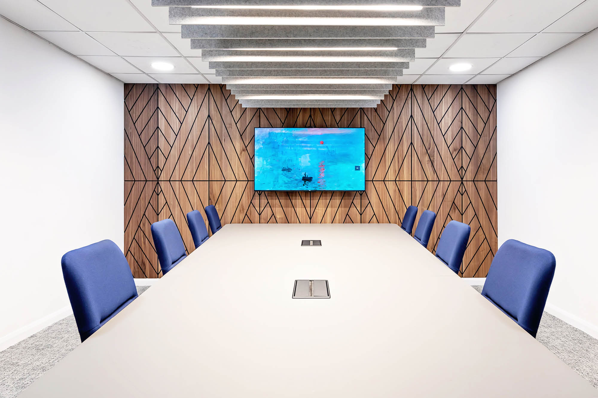 Modern conference room with a long table surrounded by eight blue chairs. A flat-screen TV is mounted on a wood-paneled wall with a geometric design.