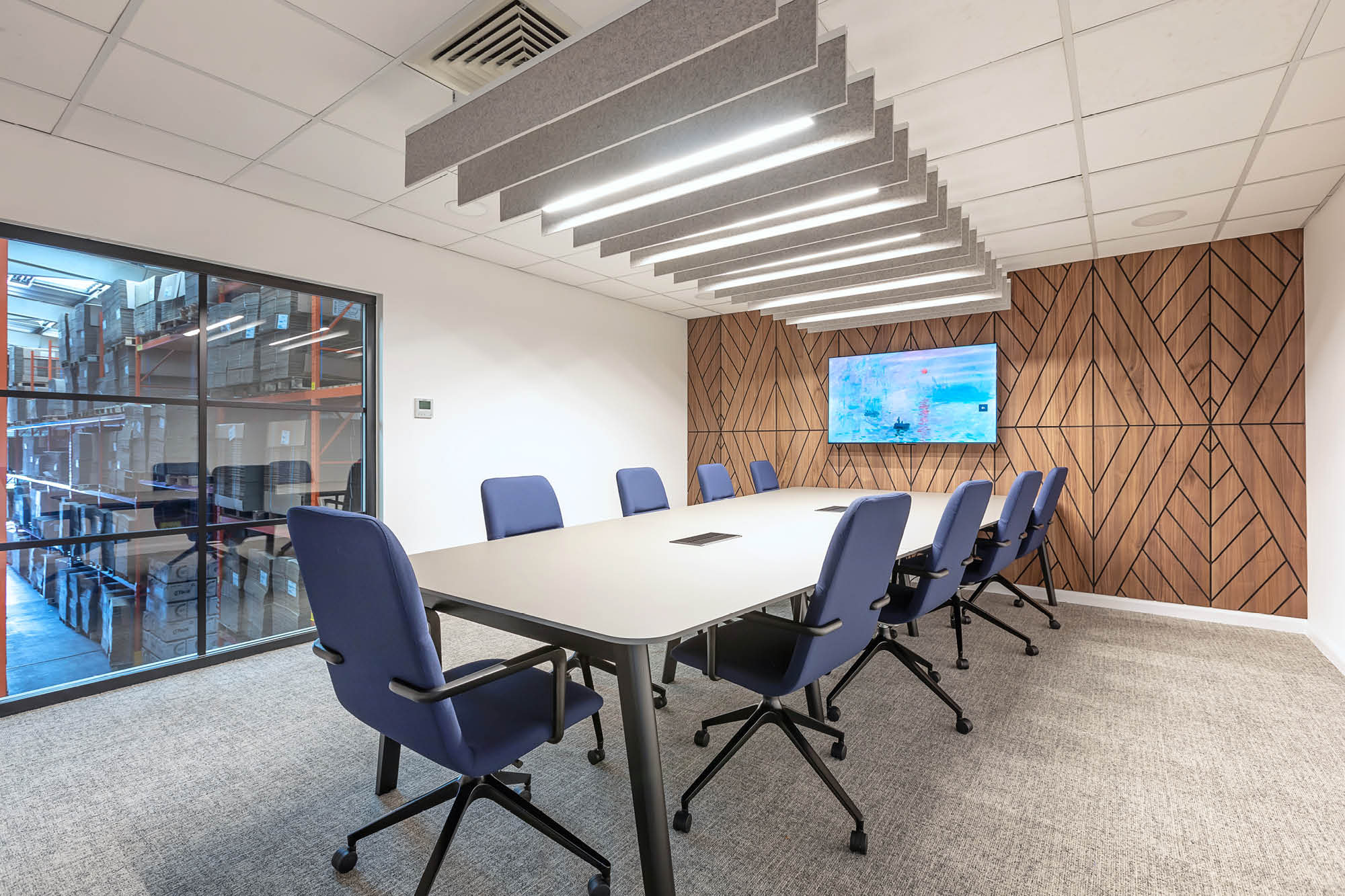 A modern conference room with large table and eight blue chairs. Overhead, there are light fixtures.