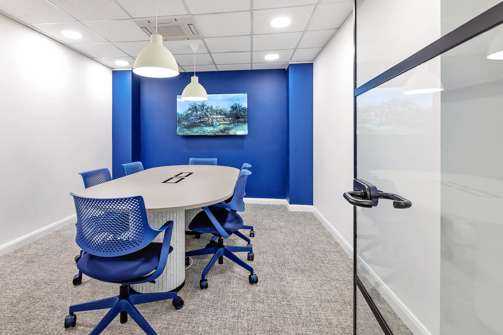 Small conference room with an oval table surrounded by six blue chairs on a gray carpet. White walls and a vibrant blue accent wall.