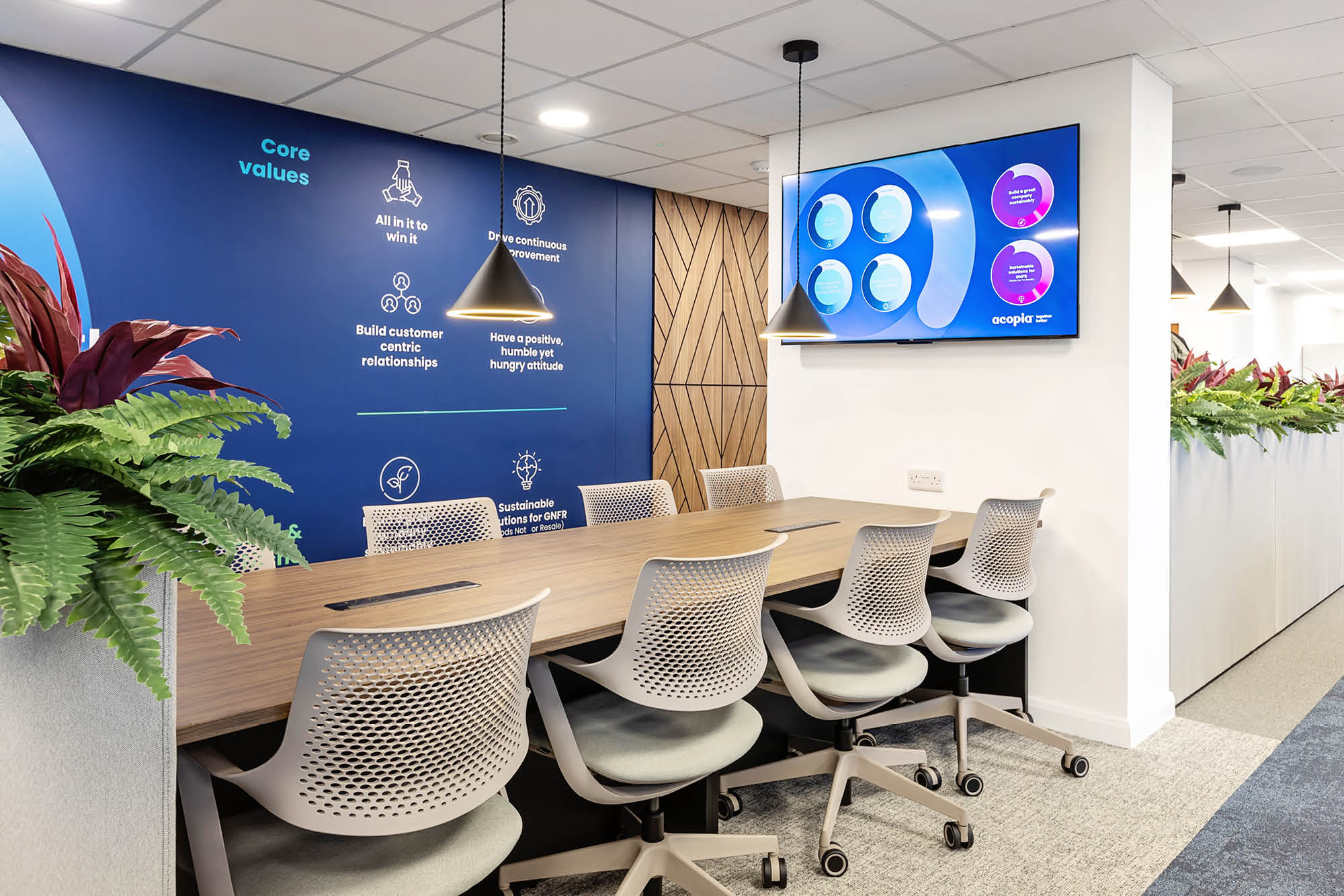 Modern conference room with a long wooden table and six ergonomic chairs. A blue accent wall displays core values.
