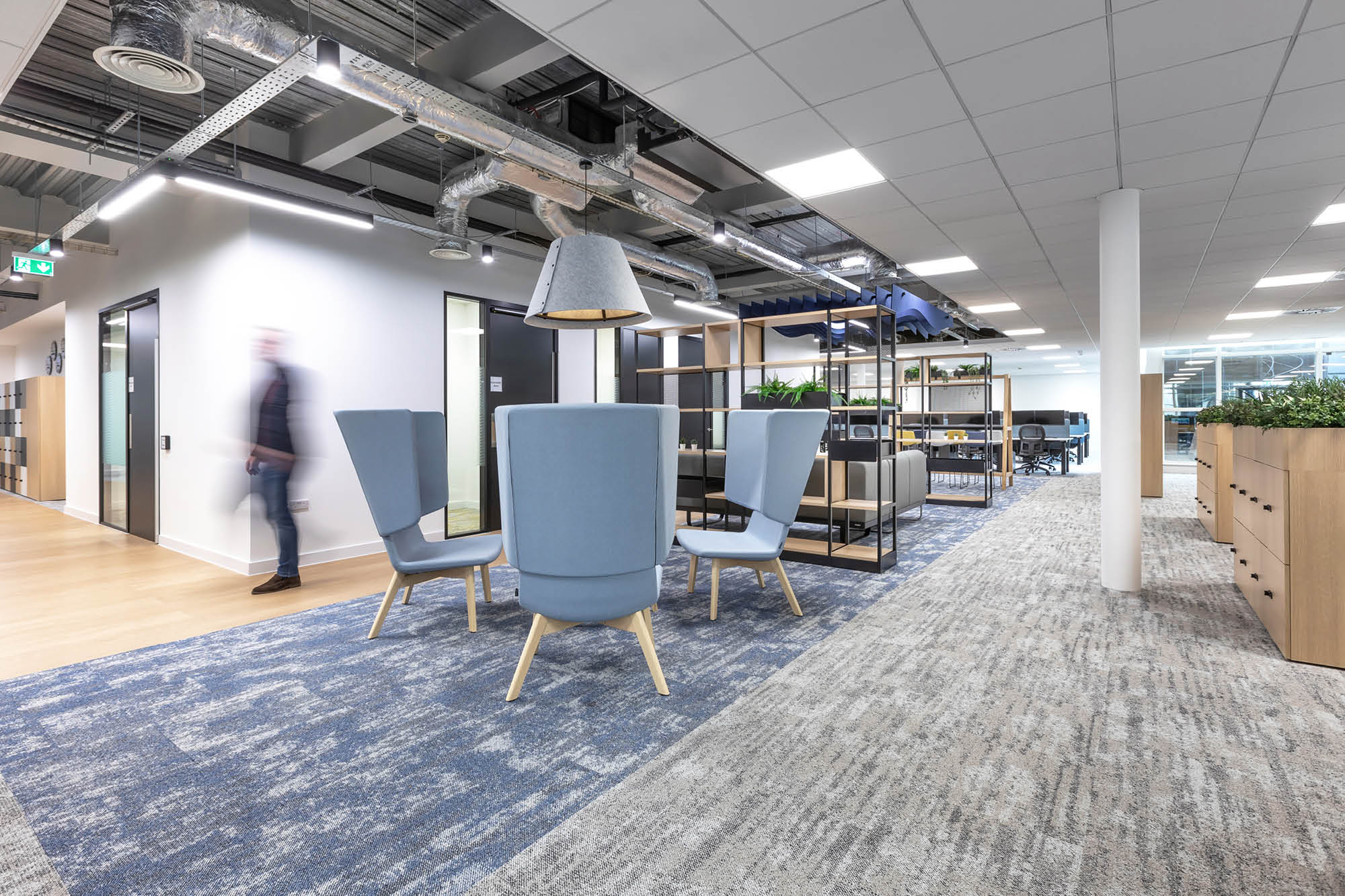 Modern office space with a blurred person walking. Features include blue lounge chairs.
