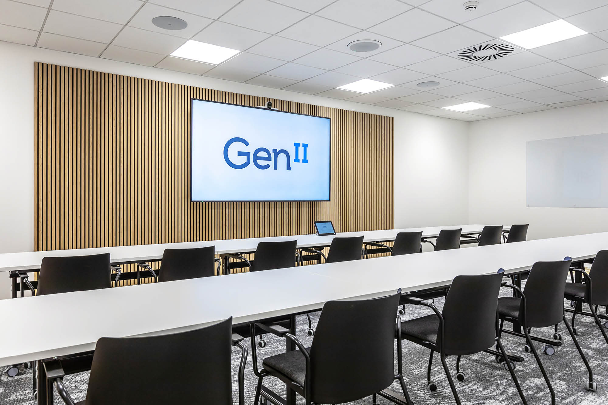 A modern conference room with rows of white tables and black chairs facing a large screen.