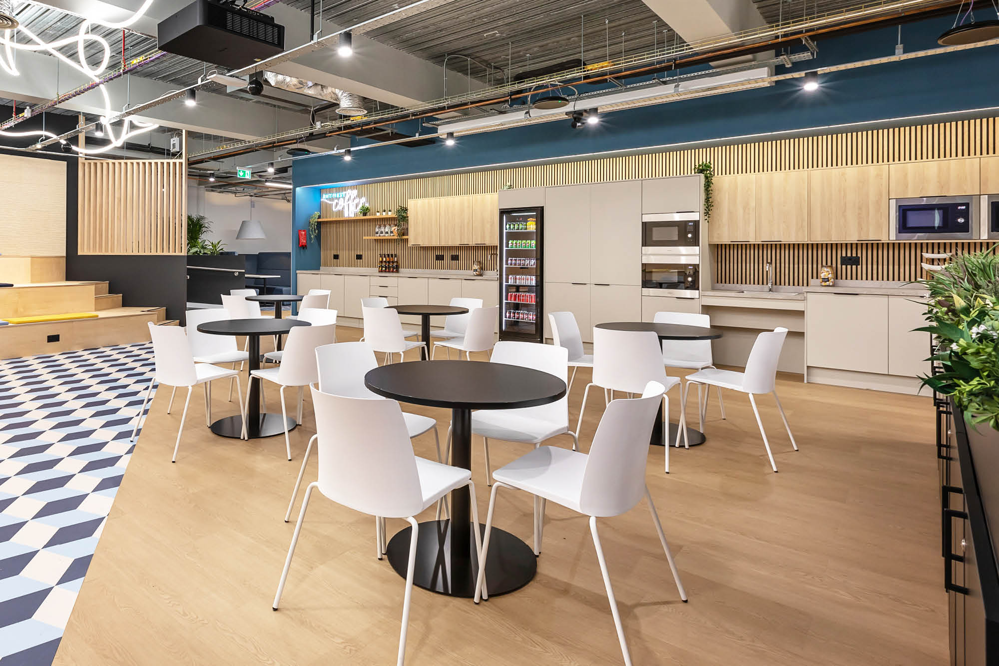 Bright modern office kitchen with white chairs and round black tables. Light wood cabinetry.