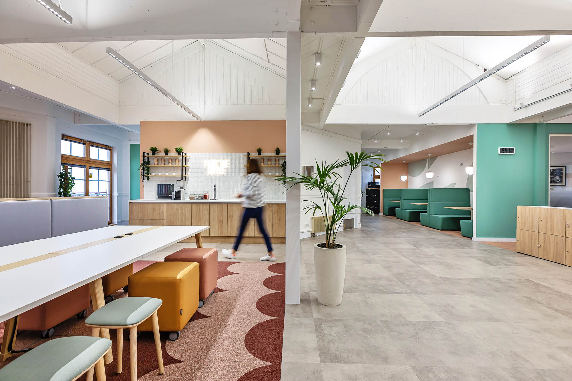 A modern office space with long white tables, stools, person walking, kitchenette.