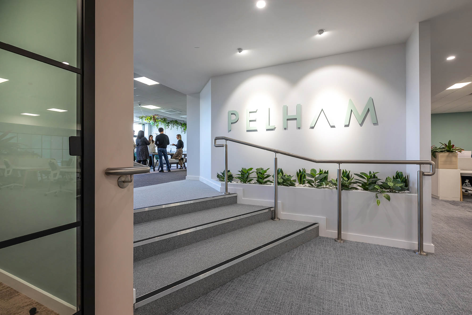 Modern office lobby with a sign reading PELHAM on wall. Steps lead to an open area of people.
