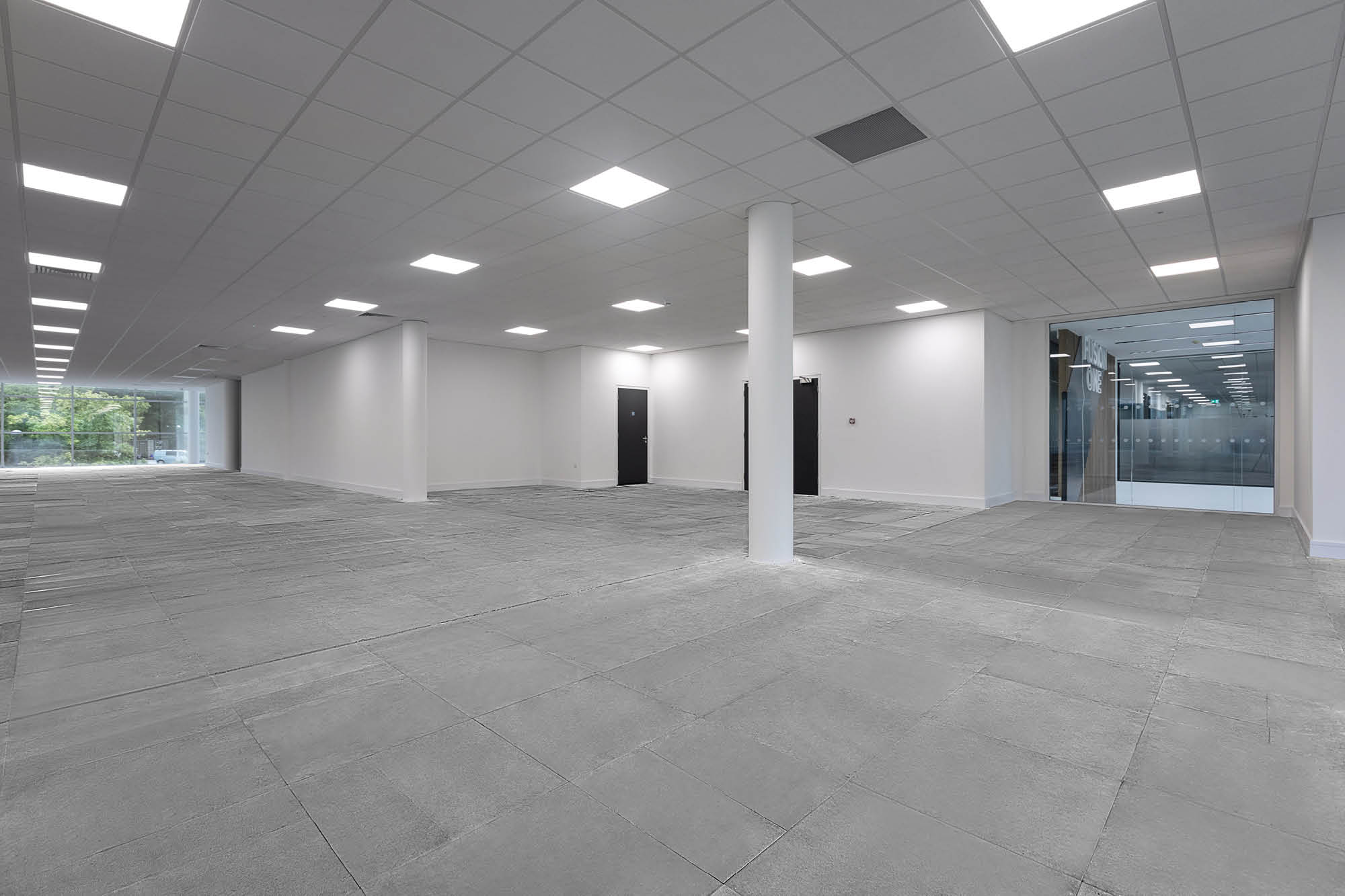 Empty office space with gray tiled floor and white walls. Ceiling is made of white panels with evenly spaced fluorescent lights.