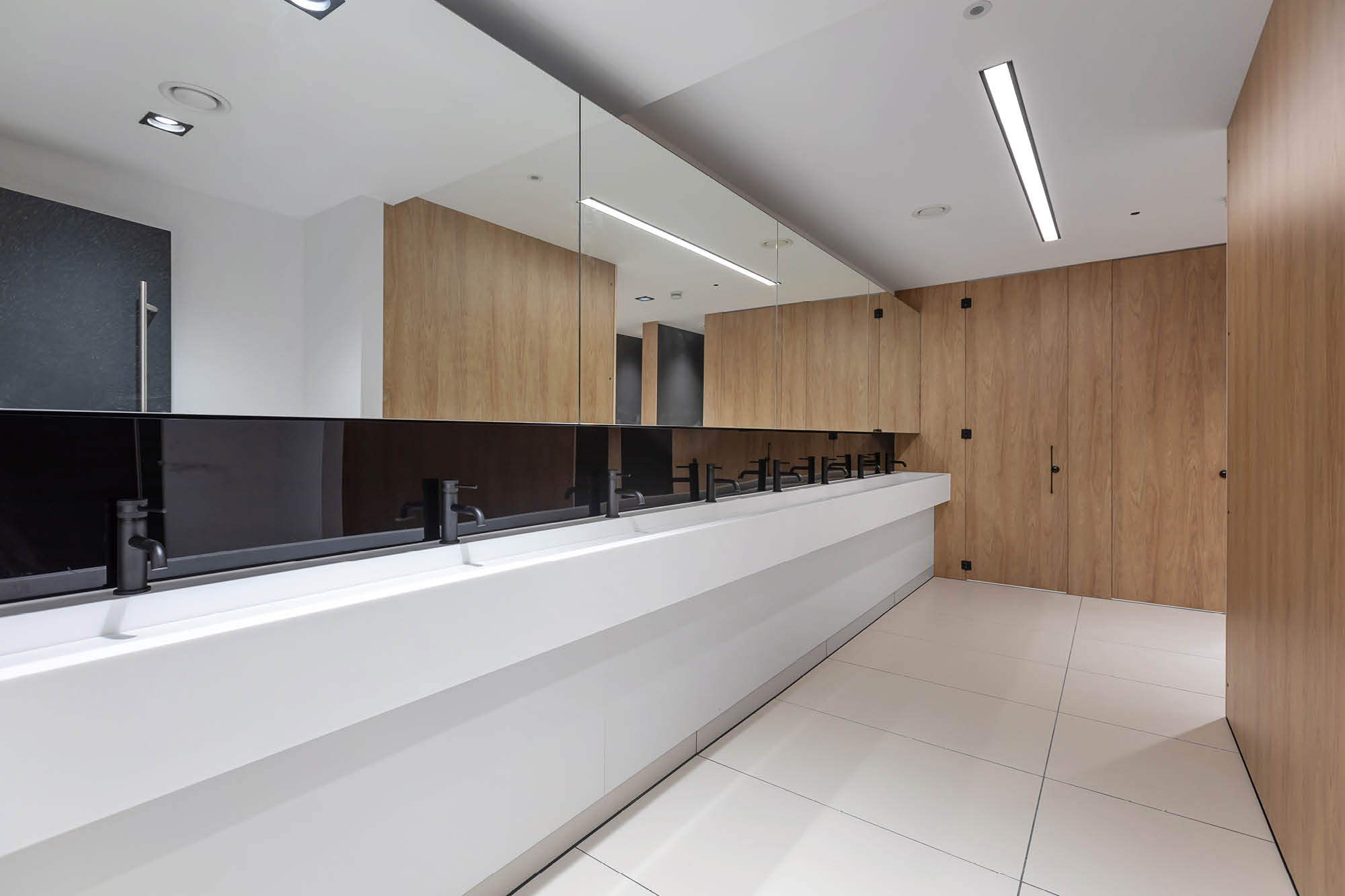 A modern public restroom with a long white counter and several black faucets, large mirrors, and wooden paneling on the walls.