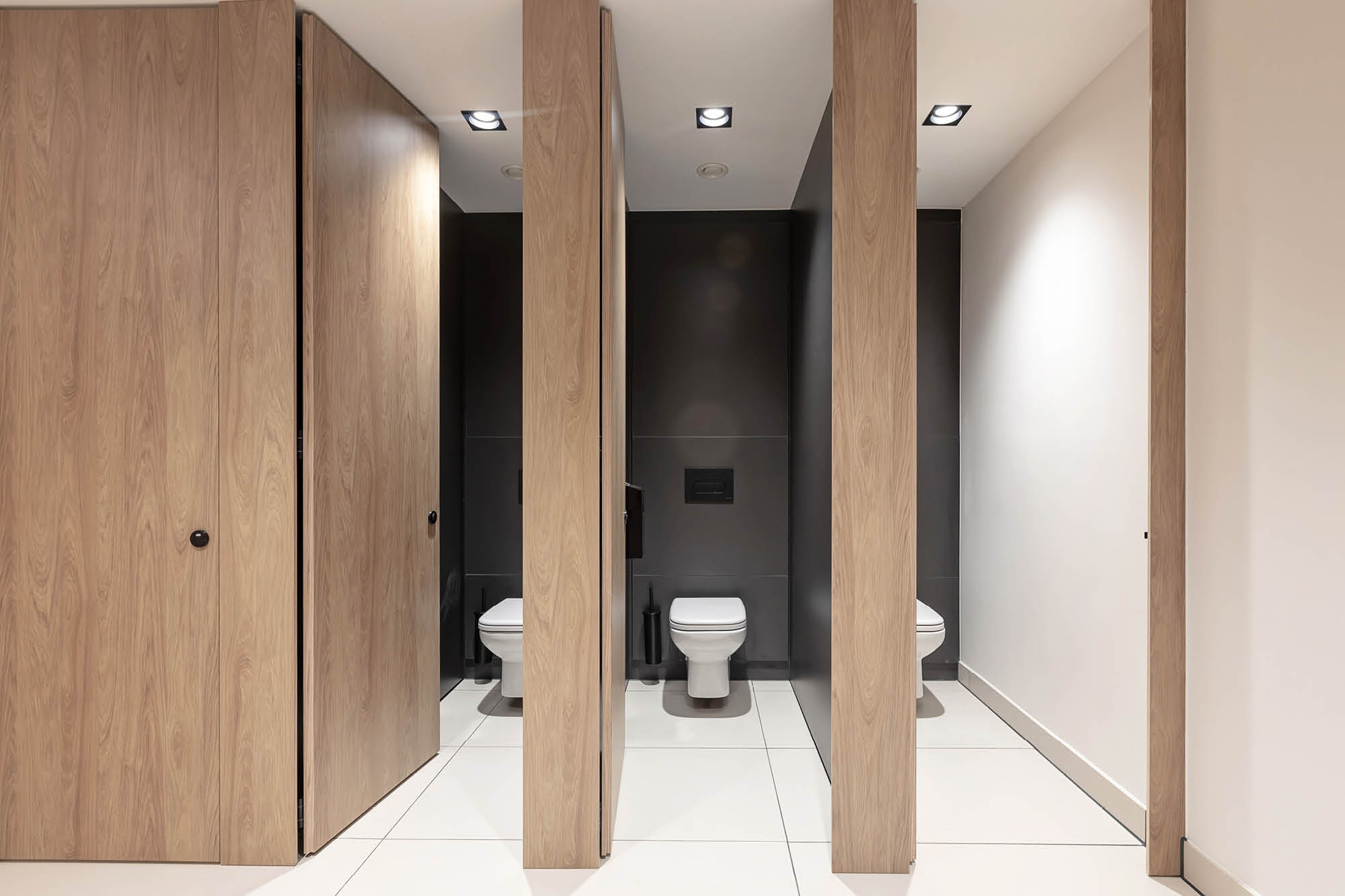 A modern public restroom with three stalls. Each stall has a wooden door, and two doors are open revealing white toilets.