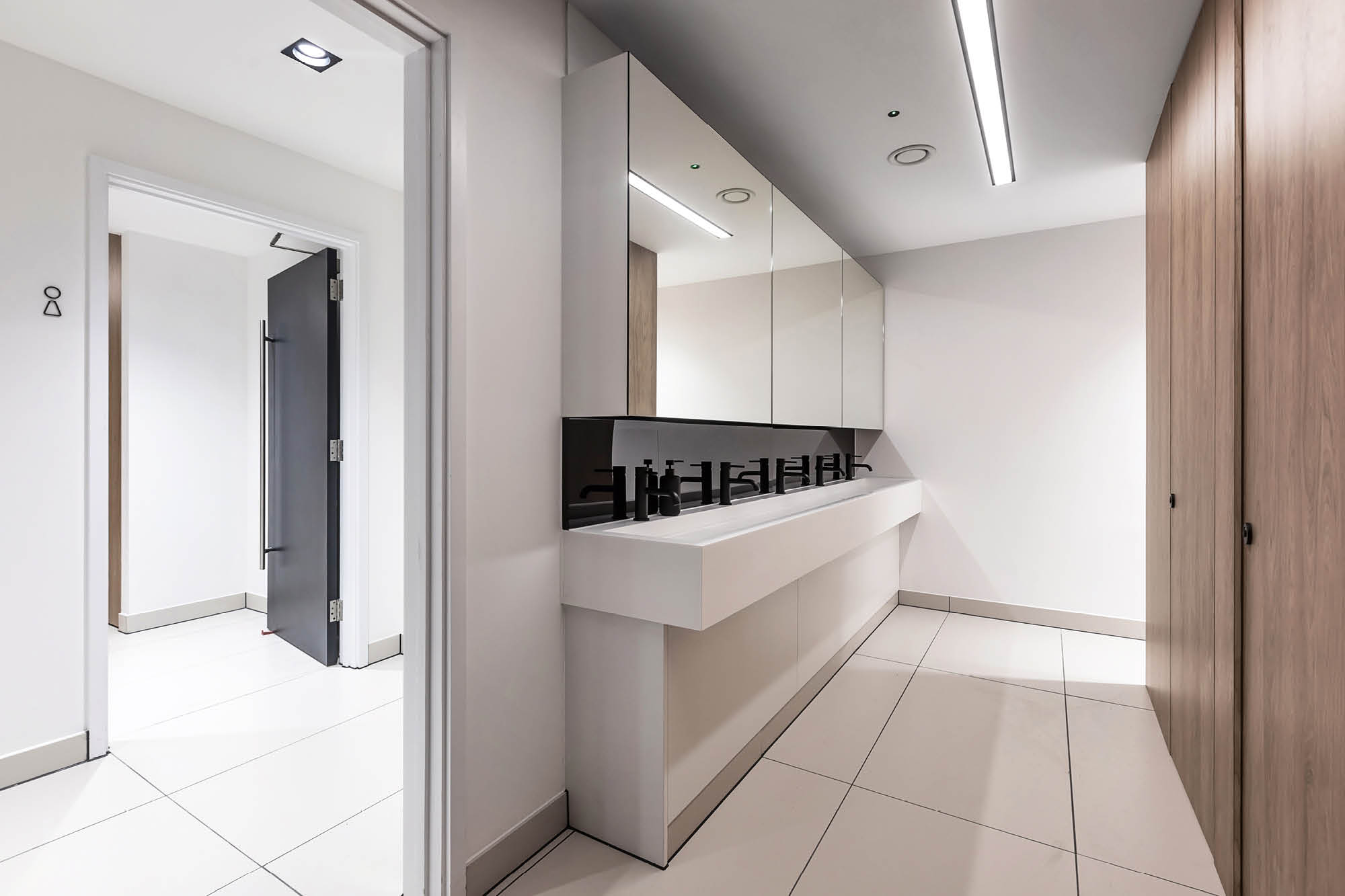 A modern restroom with white tiled floors and walls. It features a long, sleek white sink with multiple faucets and mirrors above.