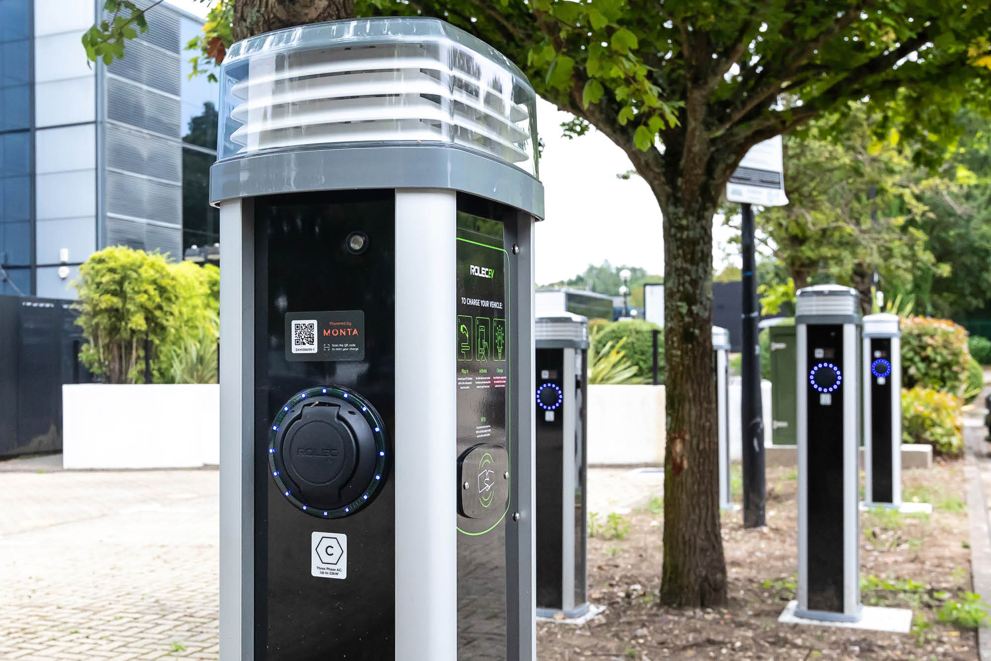 Electric vehicle charging stations lined up under a tree in an outdoor setting. Each station has a digital screen and circular LED lights.
