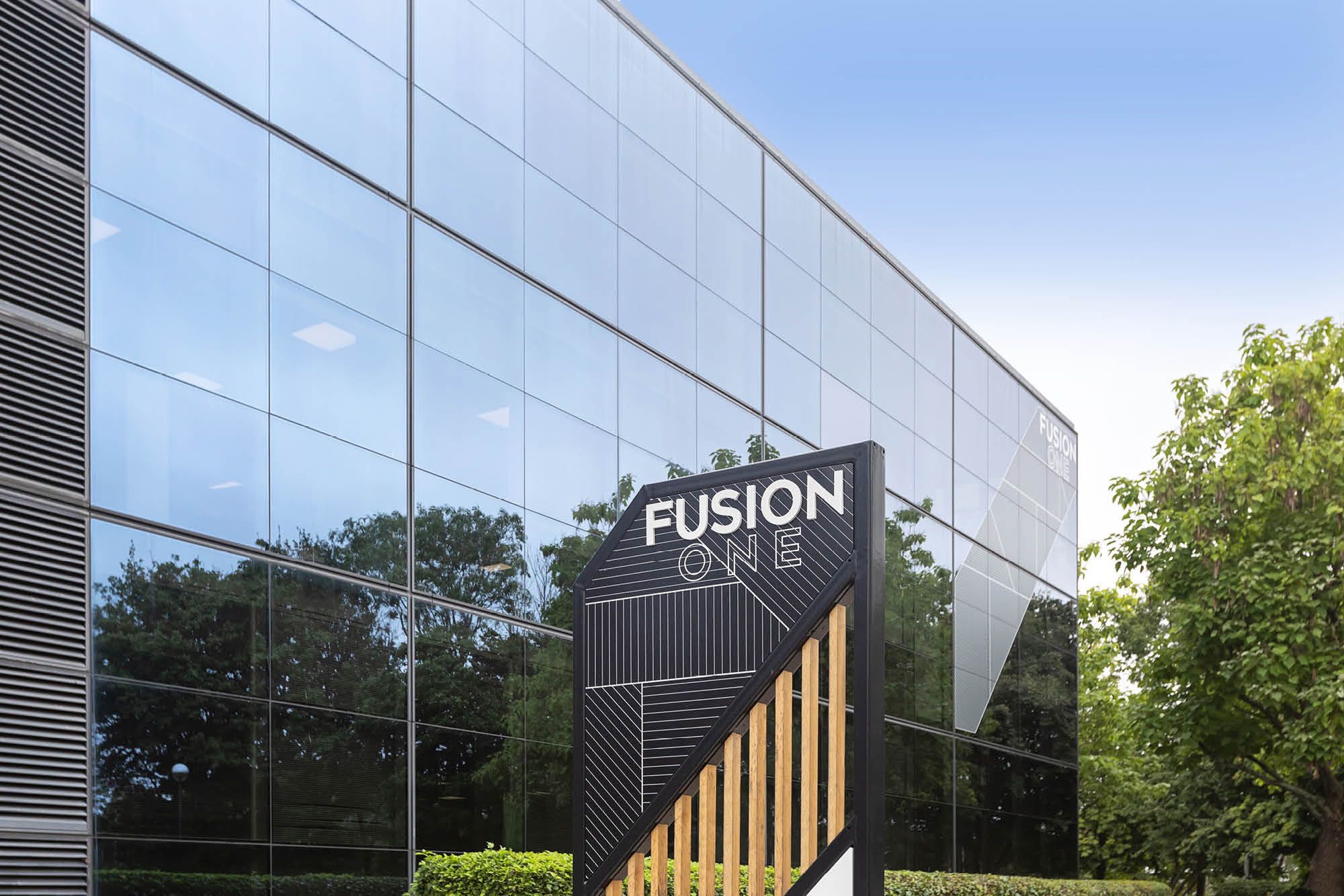 Modern glass office building with a sign reading Fusion One in the foreground. The building features reflective windows.