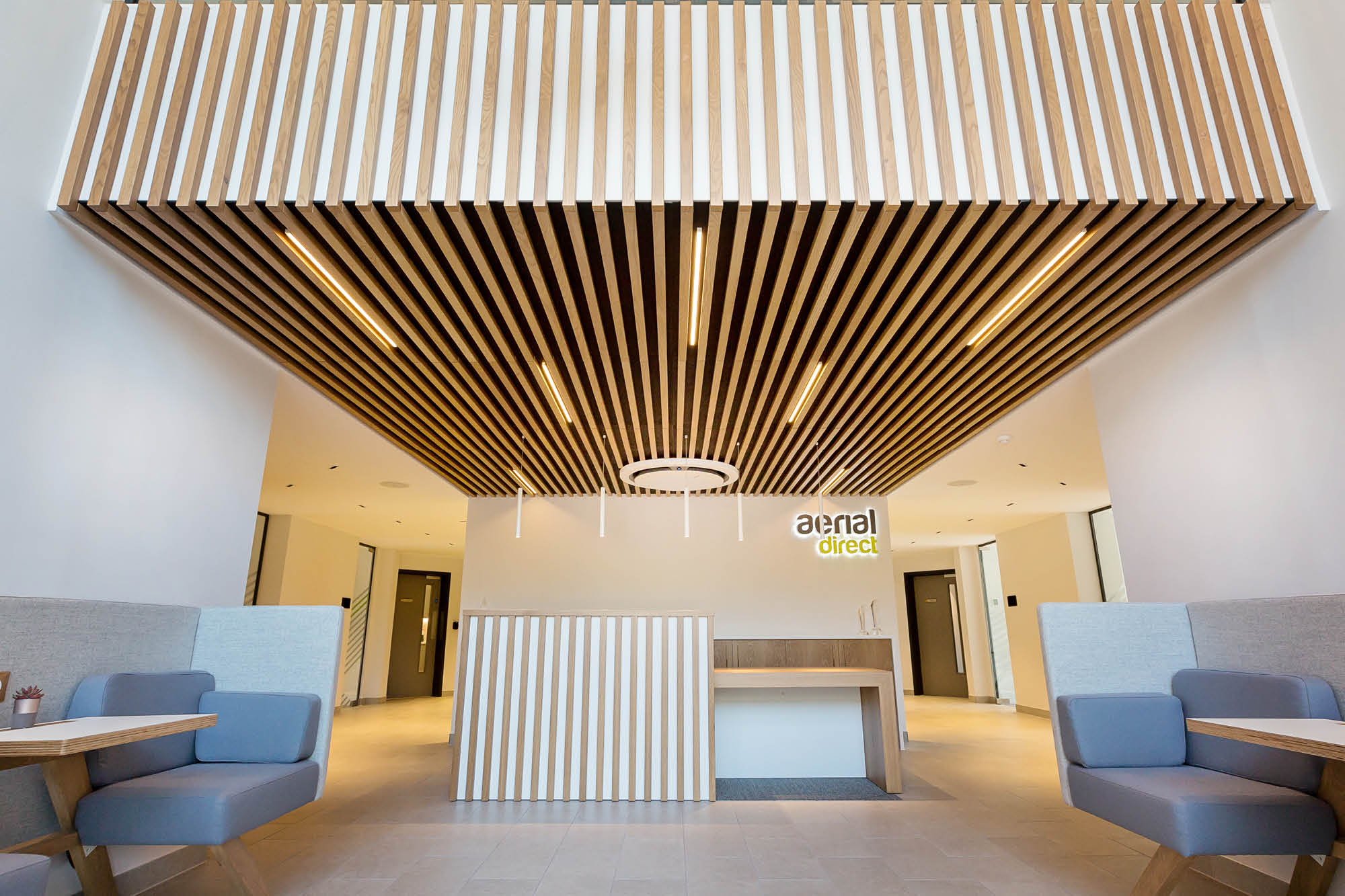 Modern office lobby with wood slat ceiling, white walls, and a simple reception desk. Blue seating booths line the sides.