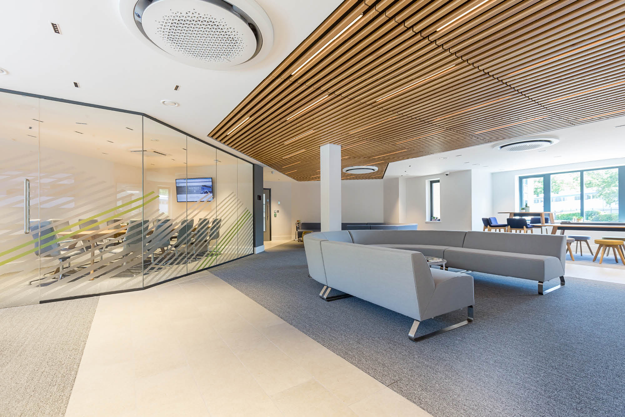 Modern office space featuring a glass-walled conference room with a table and chairs on the left. The area has a high ceiling.