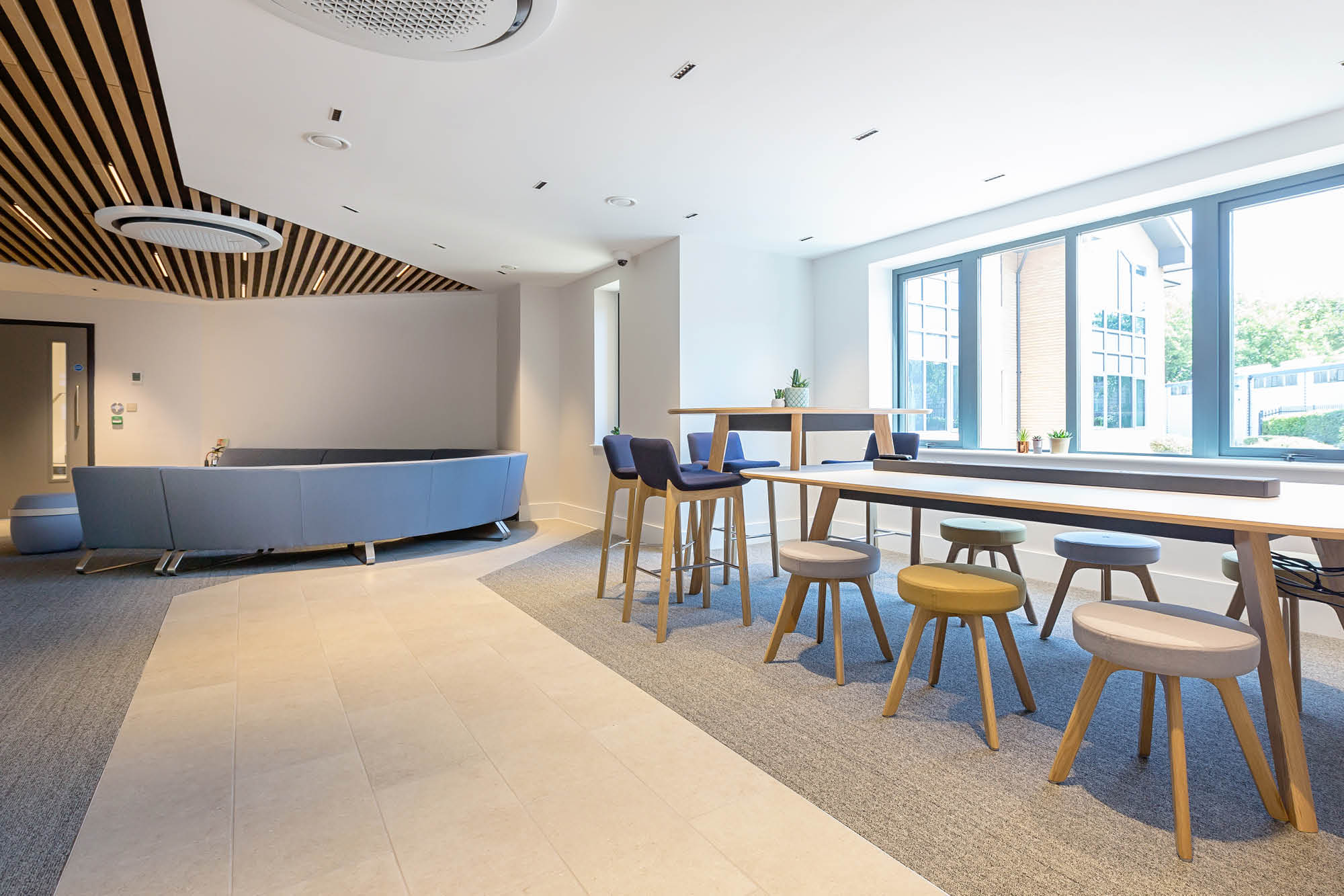 Modern office space with wood accents, featuring a round blue sofa, bar-height tables with stools, large windows letting in natural light.
