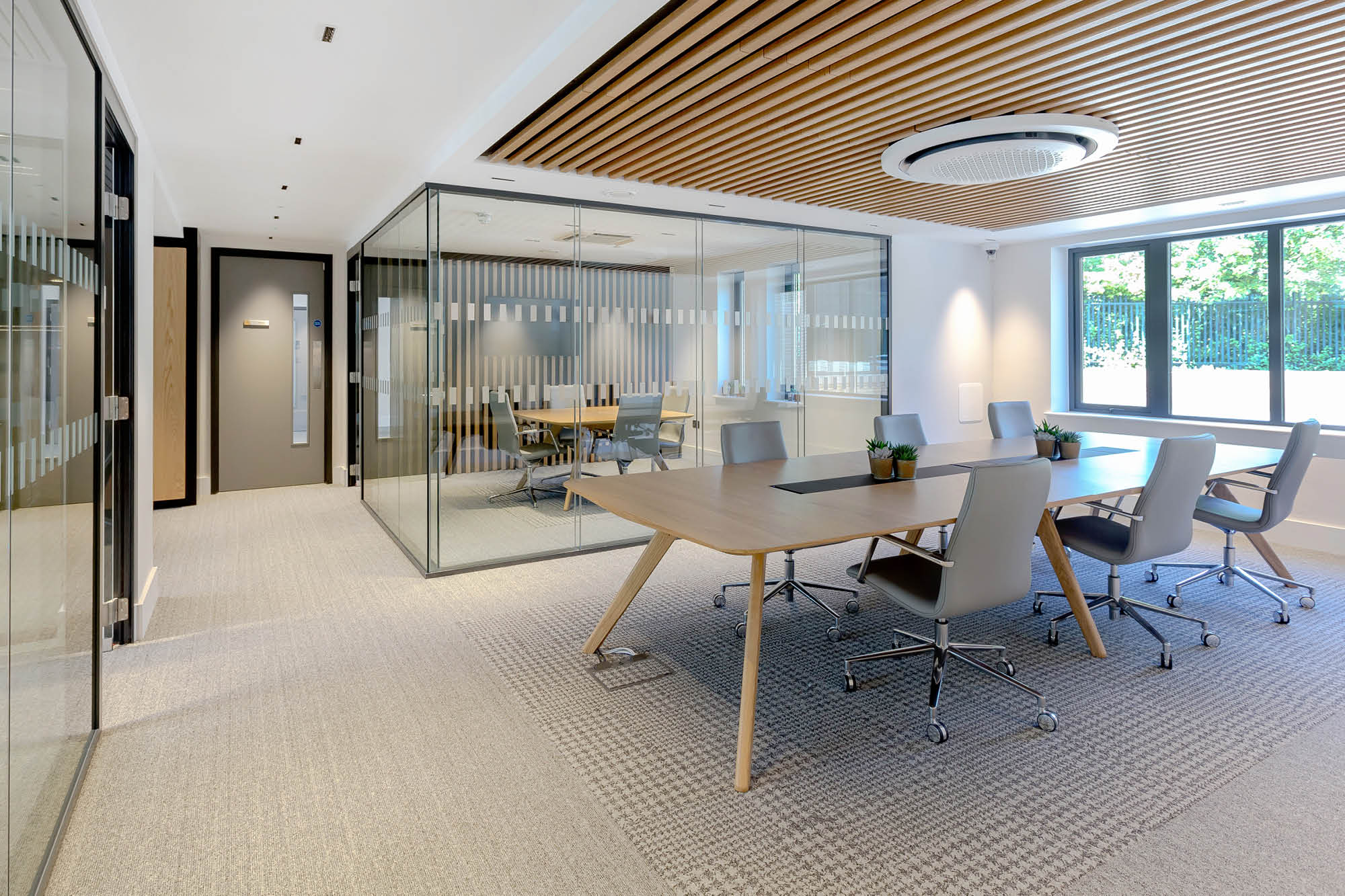 Modern office space with glass-walled meeting rooms. The main area has a large wooden table surrounded by chairs on a light carpet.
