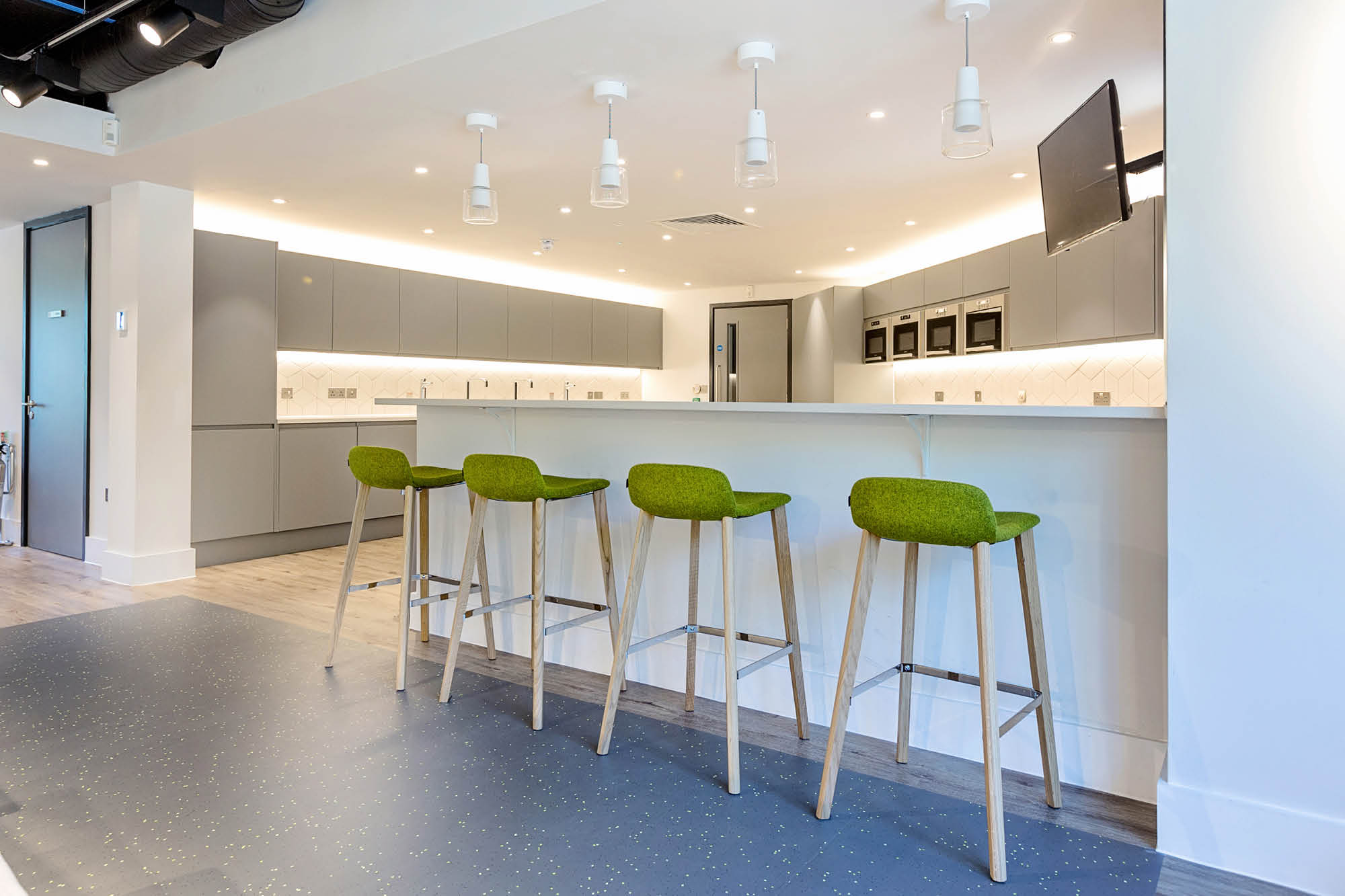 A modern kitchen area with a long white counter and four green bar stools. The space features grey cabinets, a wall-mounted TV.