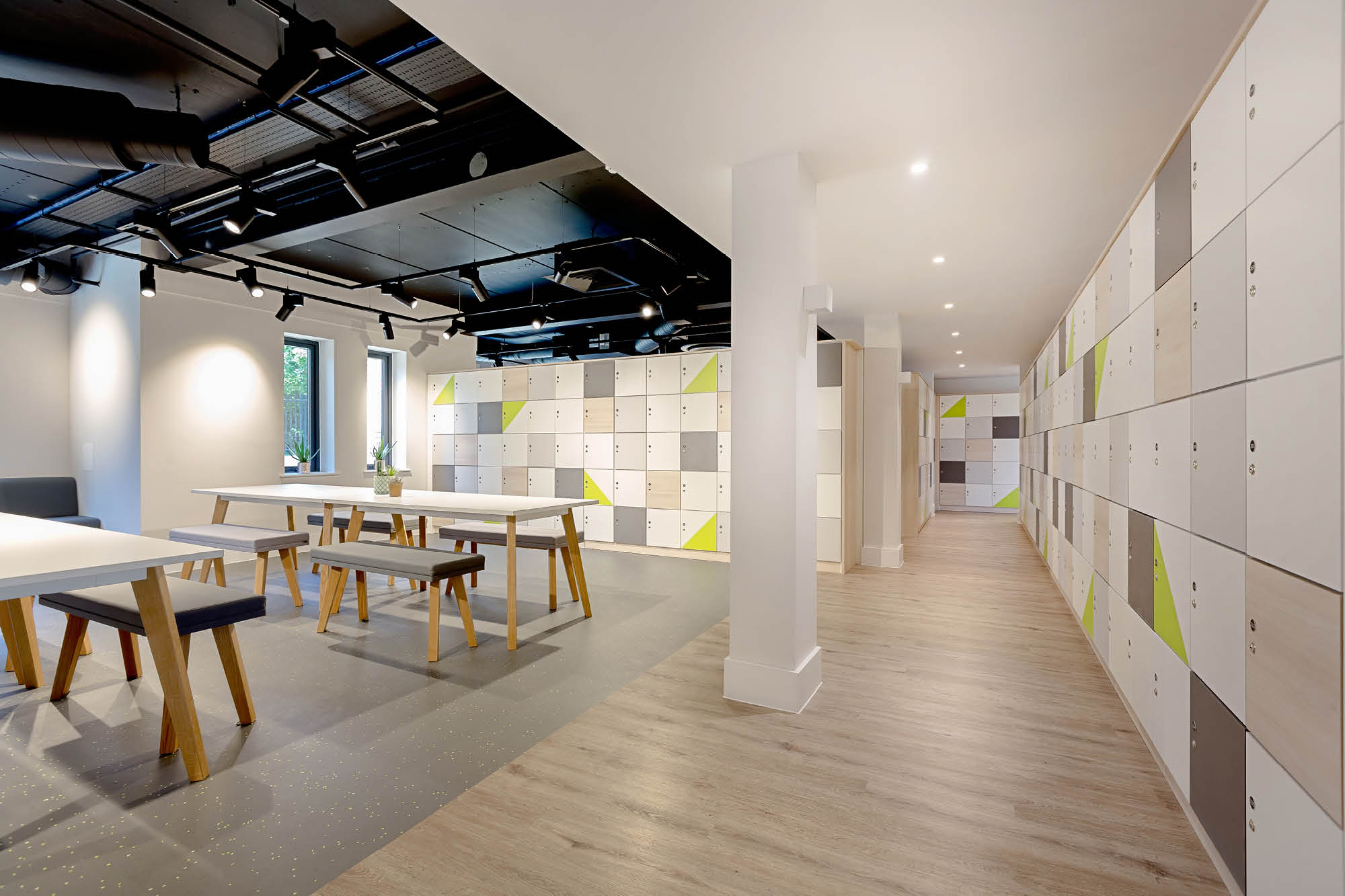 Modern interior space with tables and benches on the left and colorful lockers on the right. The room has a light wooden floor, bright lighting.