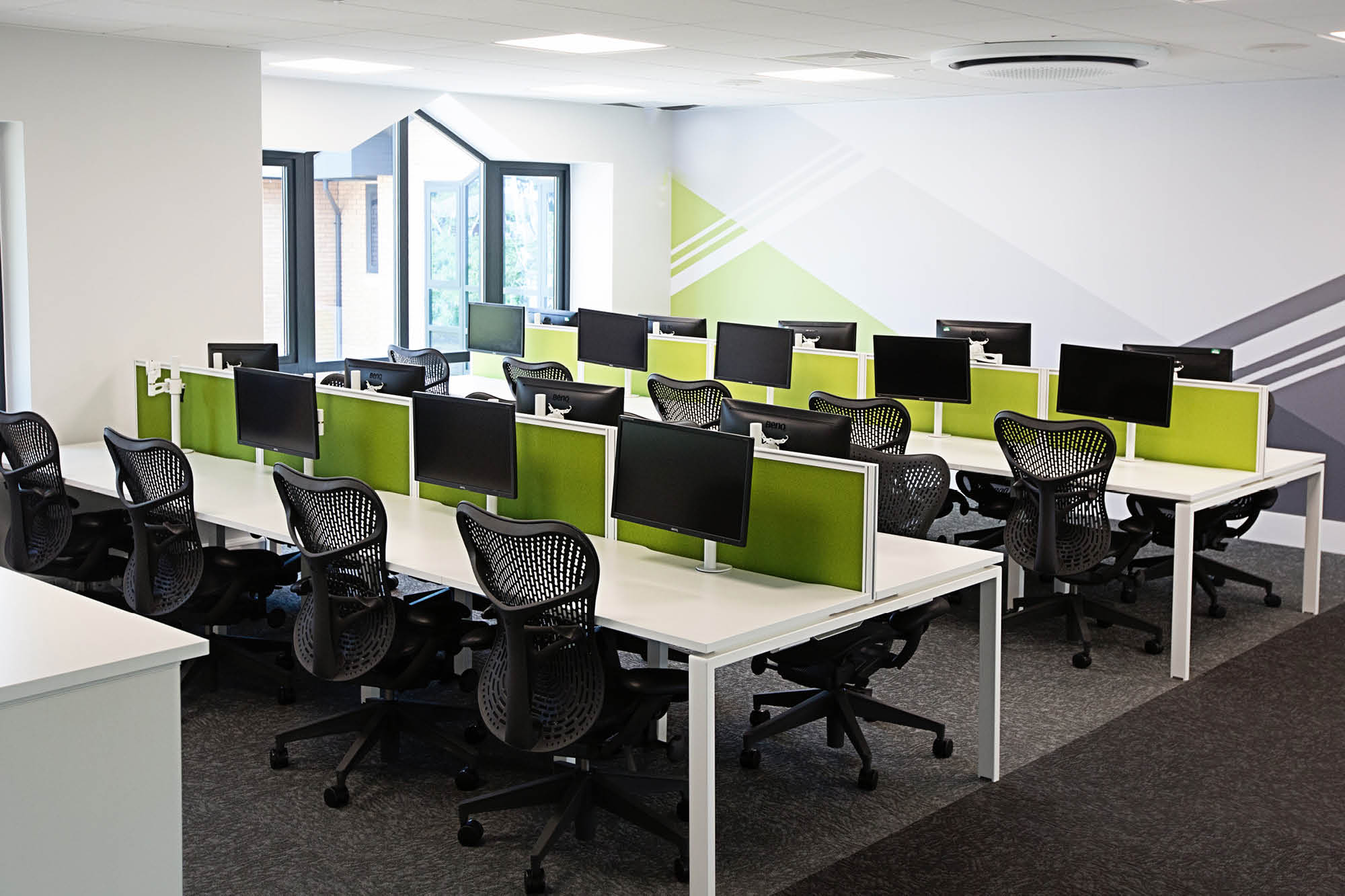 A modern office space with two rows of workstations, each equipped with a computer monitor and ergonomic chairs.