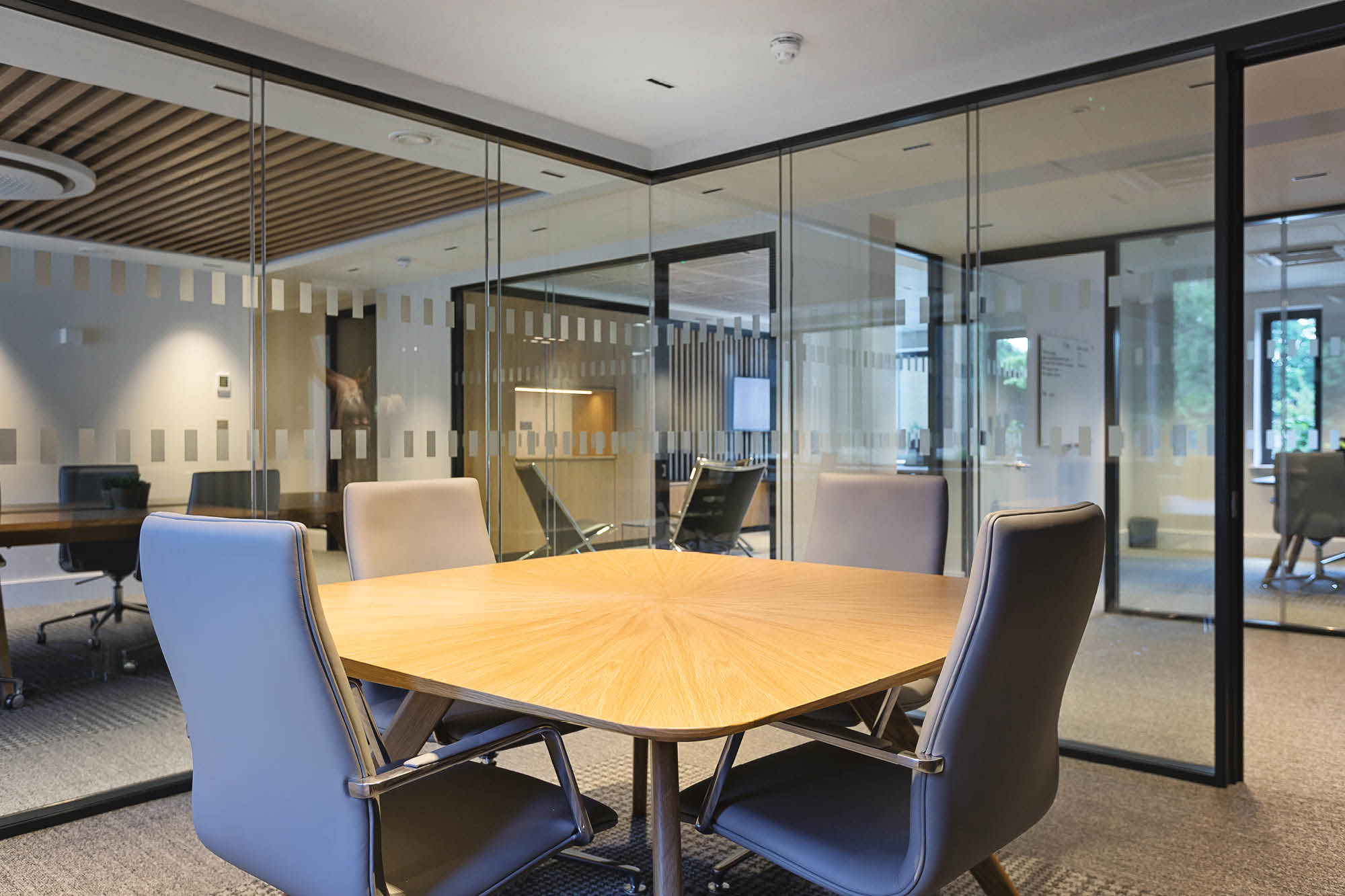 Modern office meeting room with a wooden table and four gray chairs. The room has glass walls, allowing a view into adjacent offices.