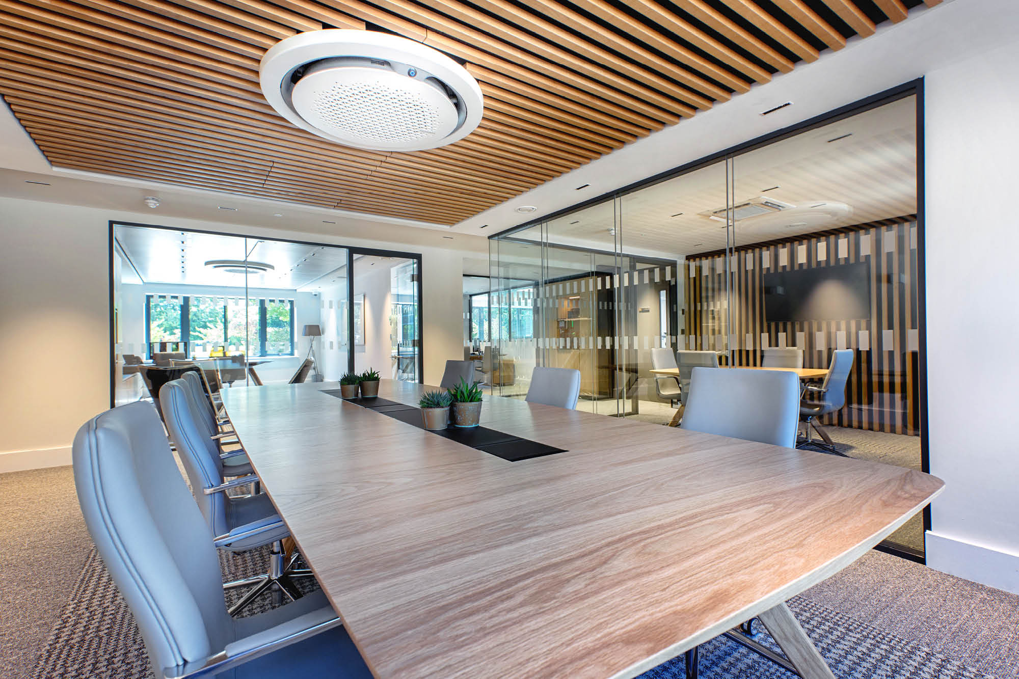 Modern conference room with a long wooden table, surrounded by gray chairs. The ceiling has wooden slats and a circular light fixture.