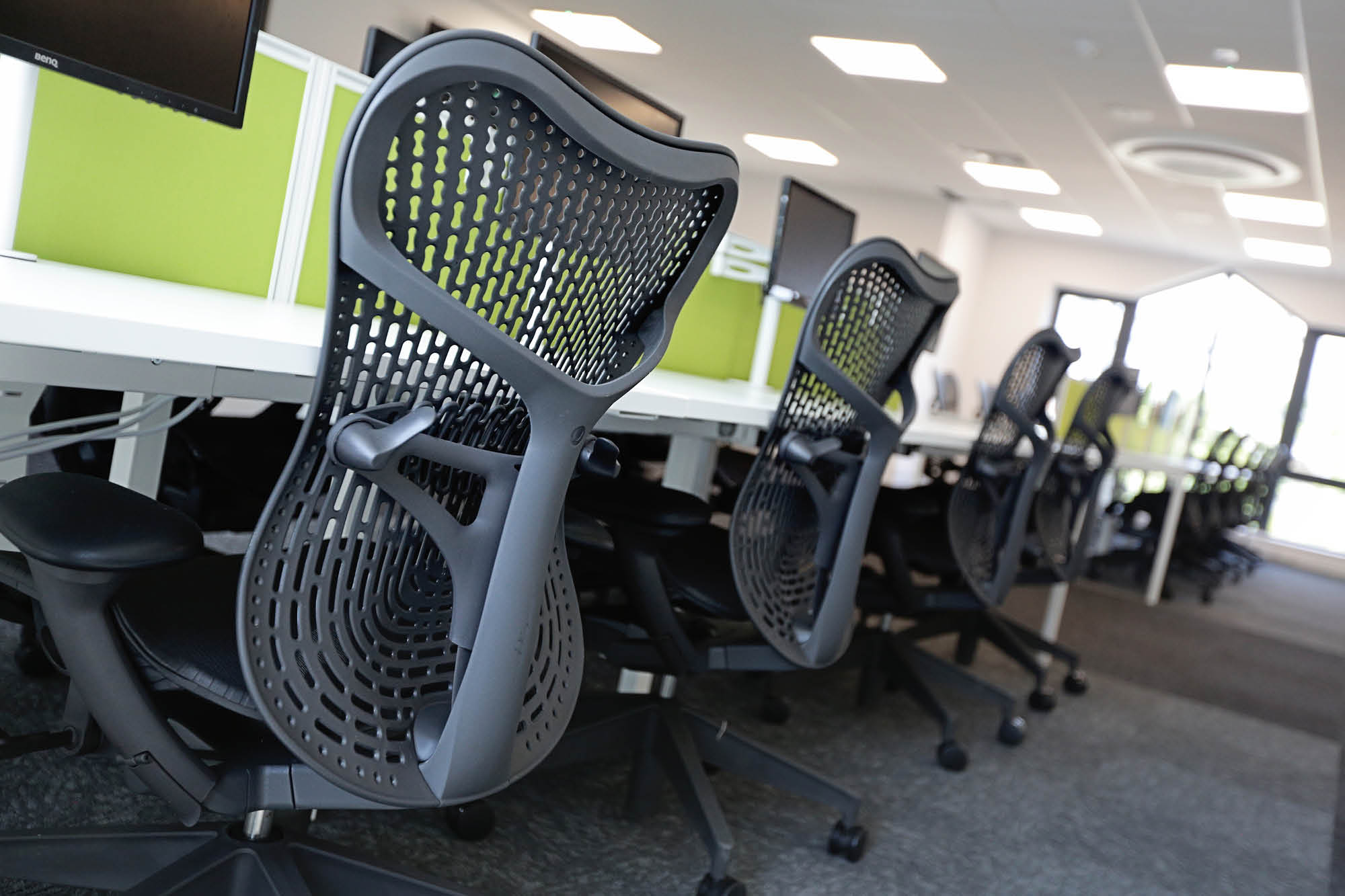 A row of modern, ergonomic office chairs with perforated backrests faces computer workstations in a bright, empty office.