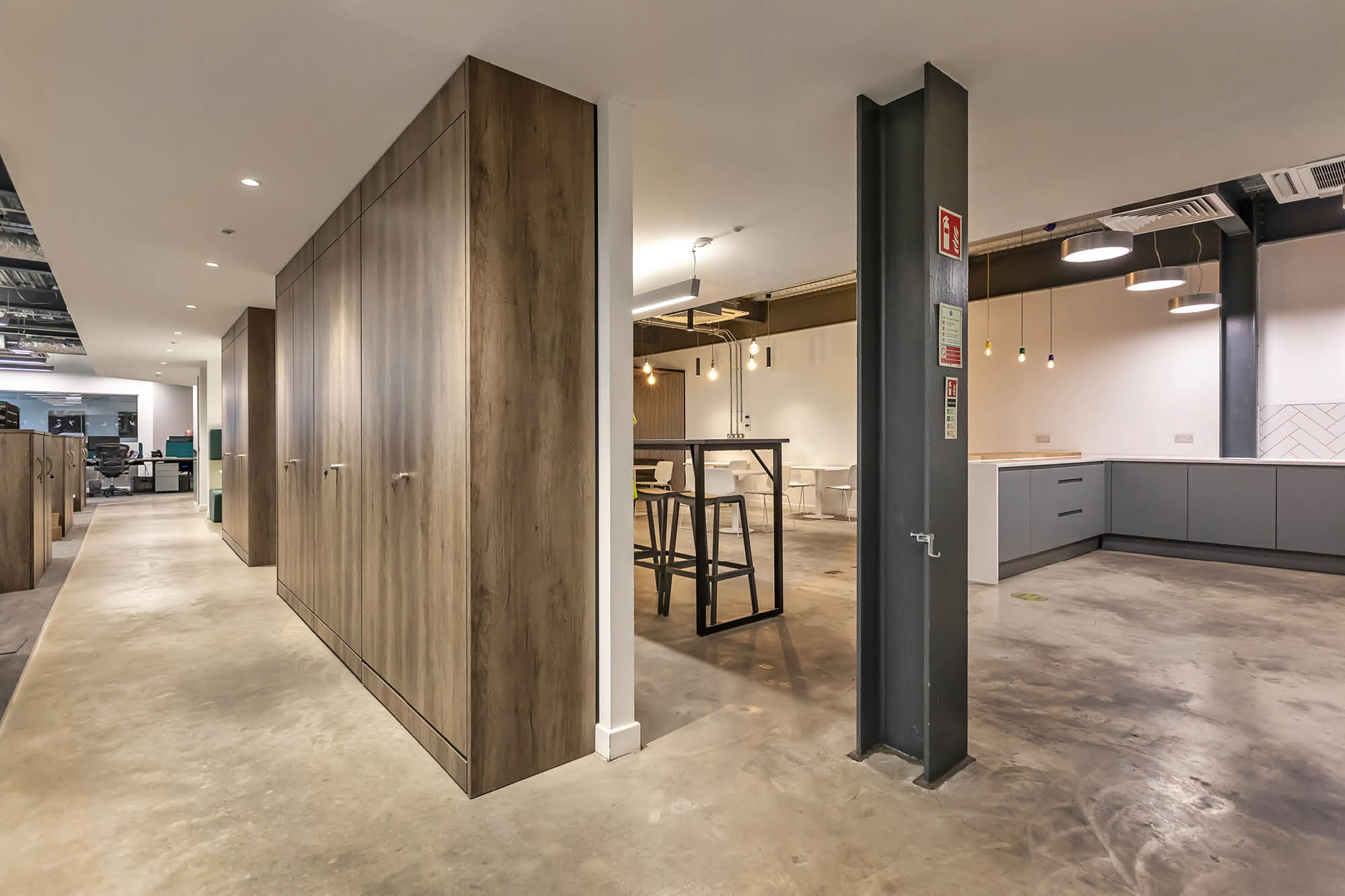 A modern office interior featuring a long hallway with wooden cabinets on the left, an open seating area with bar stools and high tables in the centre.