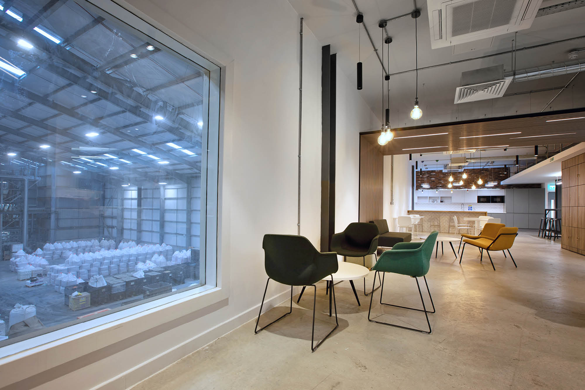 A modern office lounge with chairs and tables arranged near a large window overlooking a warehouse. The space features exposed lighting.