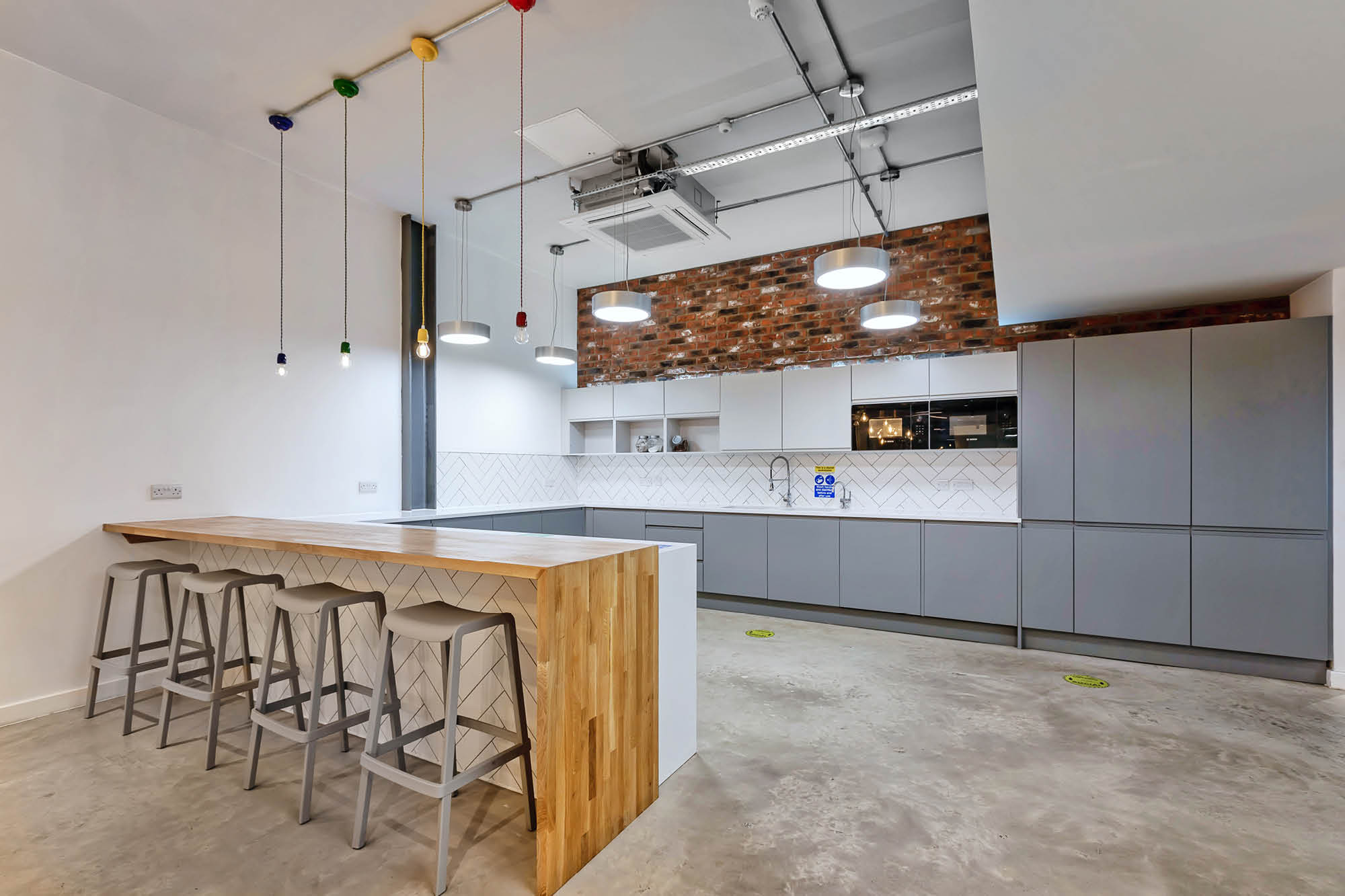 Modern kitchen with industrial design features. A wooden island has four gray bar stools. Overhead, multicolored pendant lights hang from the ceiling.