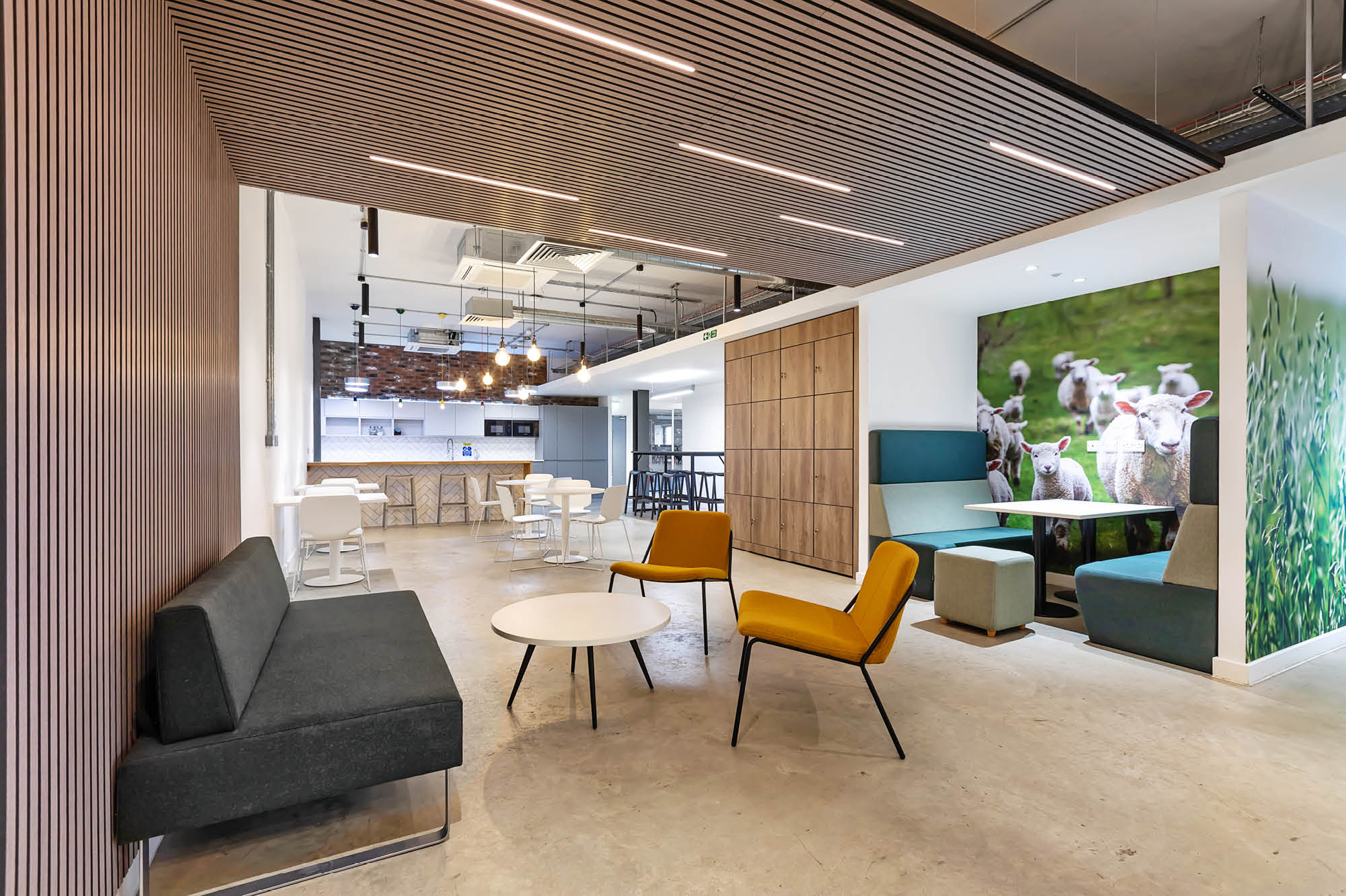 Modern office lounge with a black sofa, yellow chair, and round white tables. In the back, a kitchenette with bar stools.