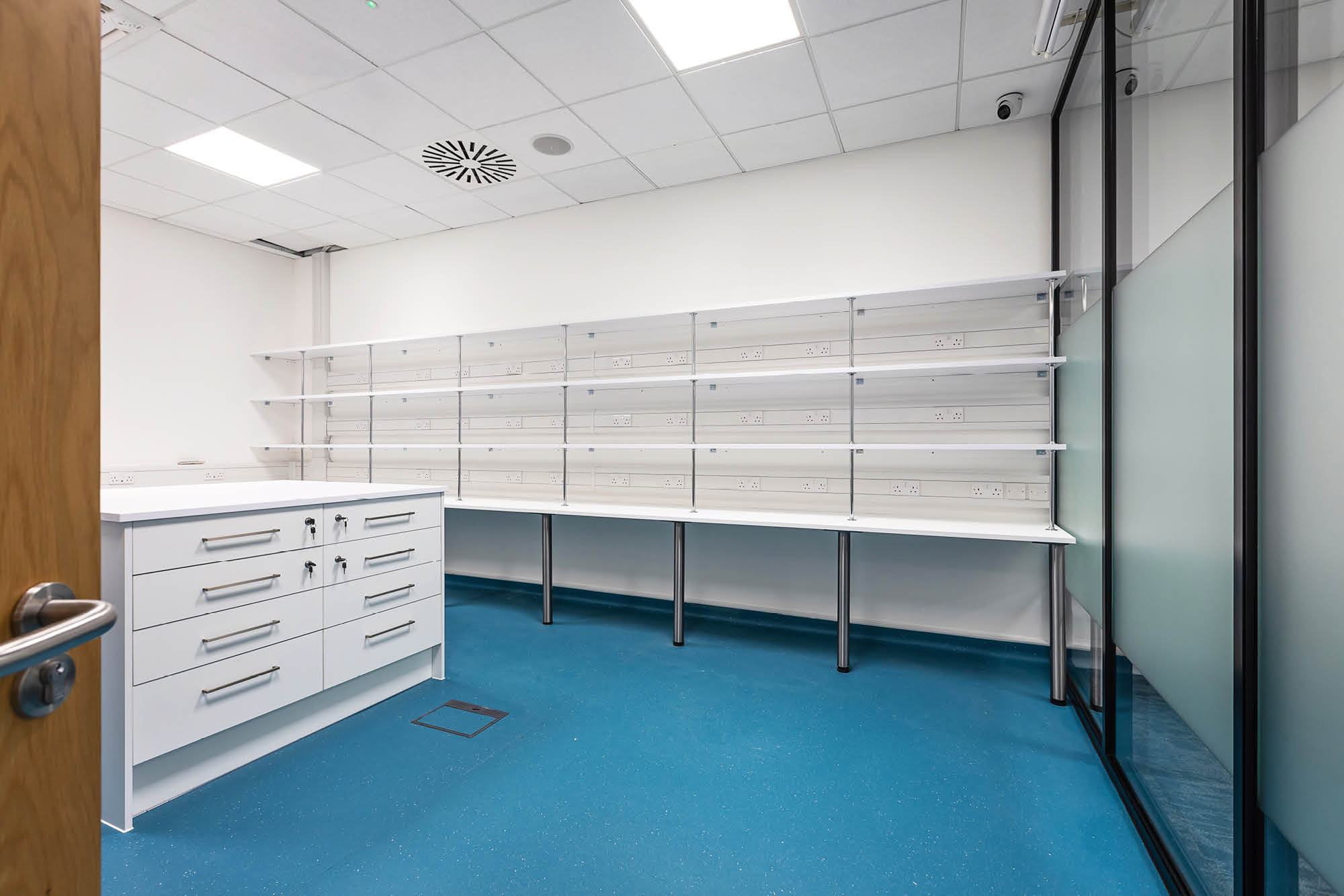 A modern, empty laboratory room with white walls and blue flooring. It features white cabinets with drawers.