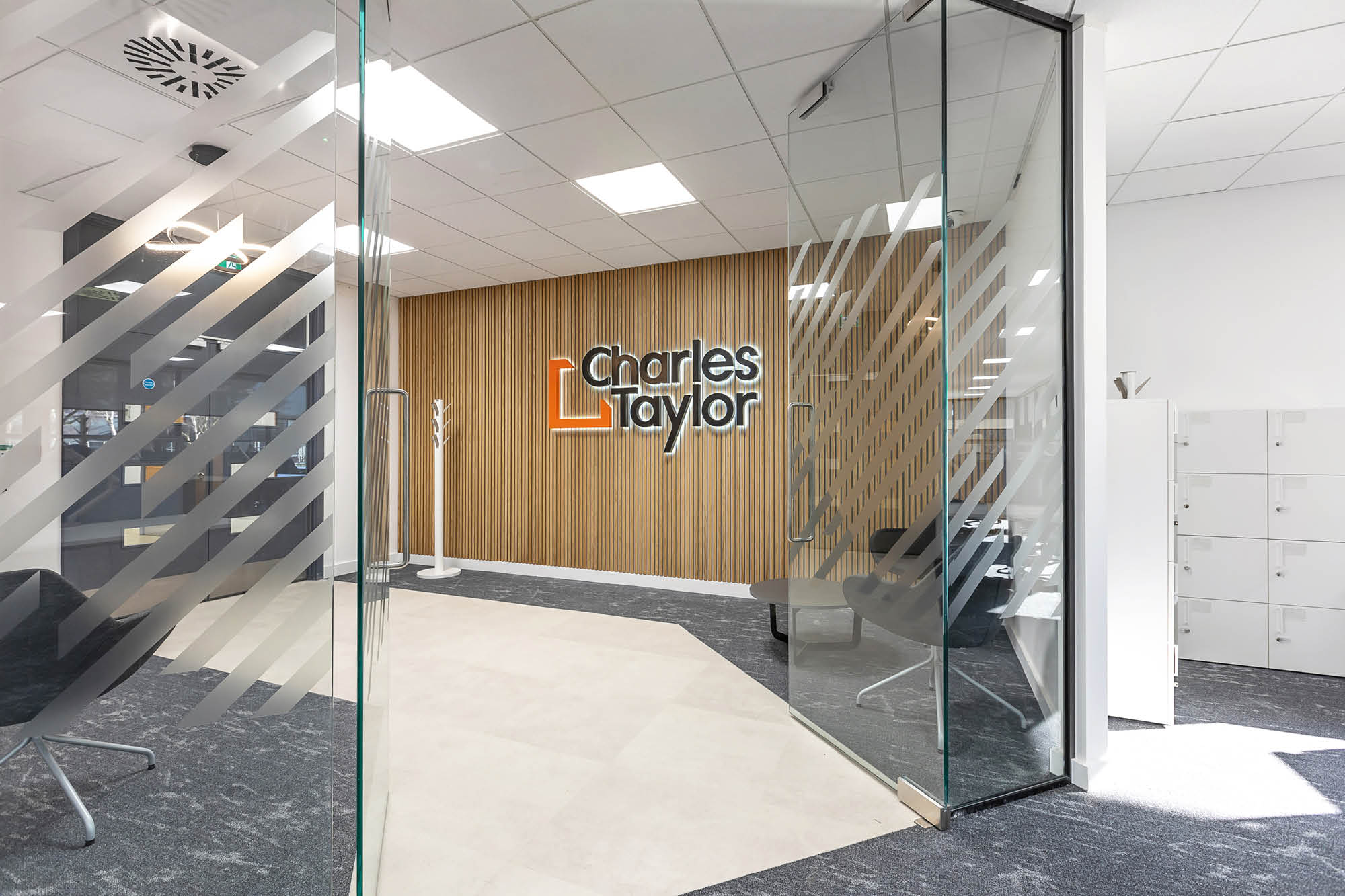 Office entrance with glass doors featuring a geometric design. Inside, a wood-paneled wall displays the logo Charles Taylor.