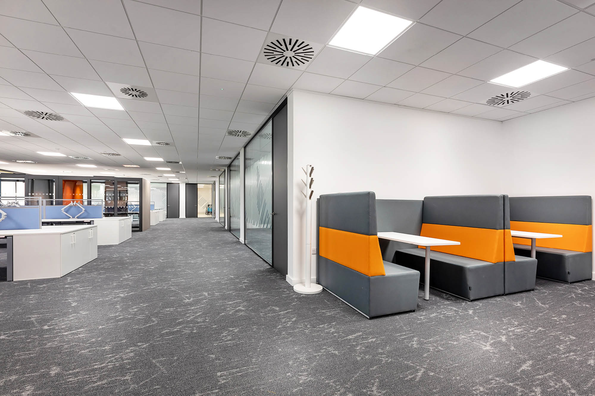 Modern office space with gray carpet, glass partition, and white ceiling tiles. Features gray booths with orange accents.
