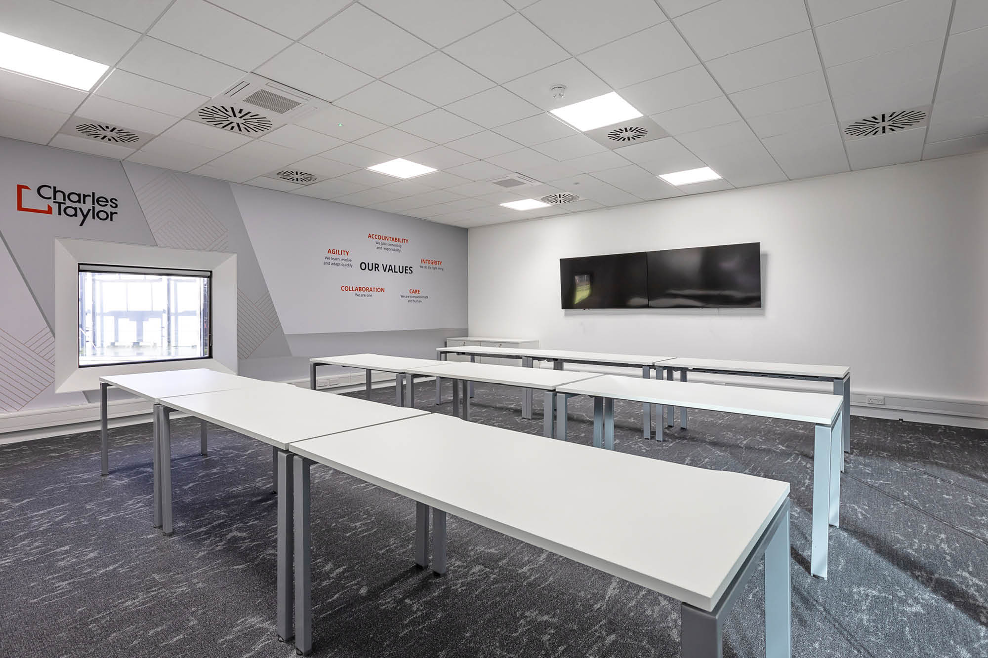 A modern classroom with white tables and a large flat-screen TV on the wall. The walls have motivational words and a company logo.