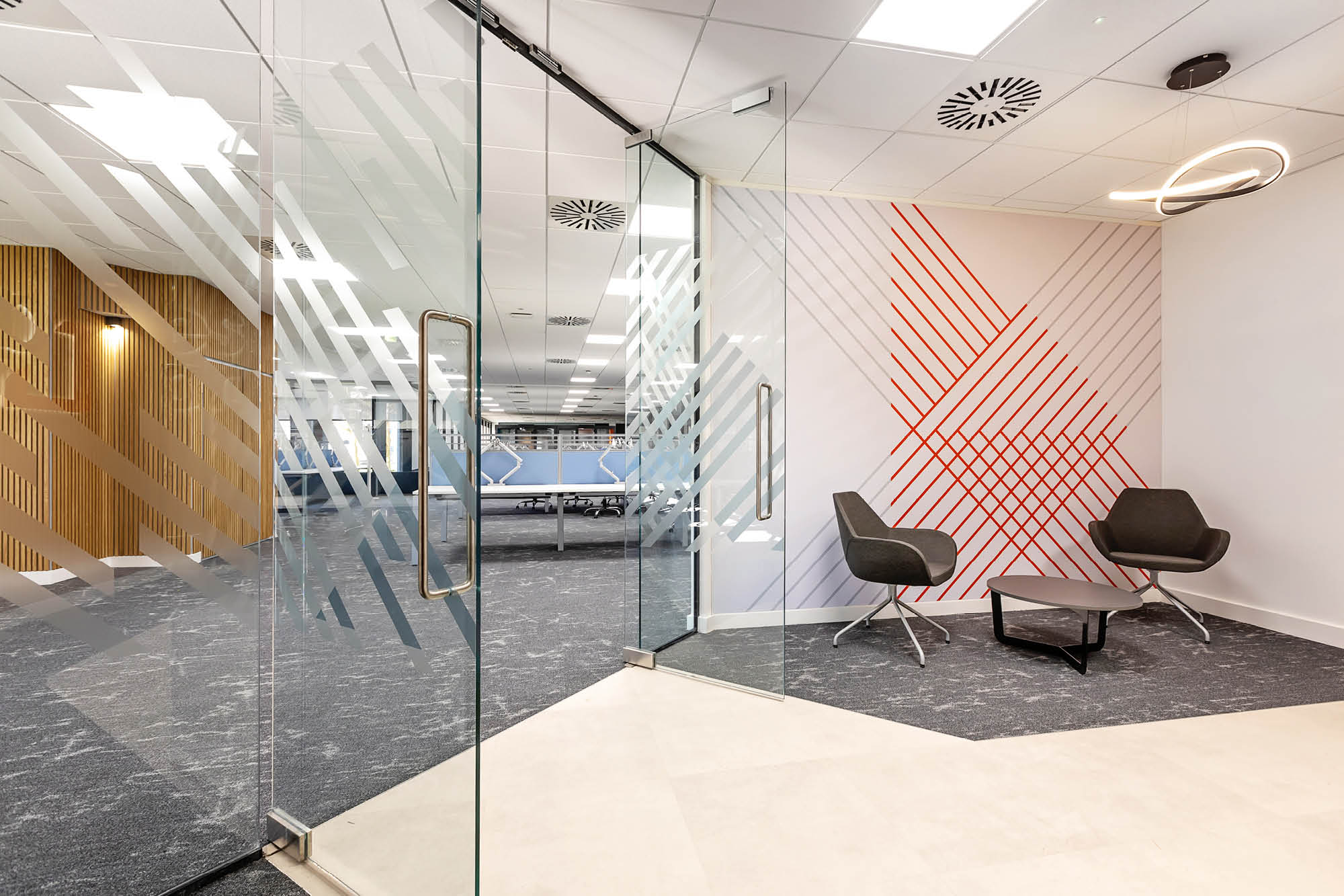 Modern office lobby with two black chairs and a small table on dark carpet. The wall has red geometric patterns.